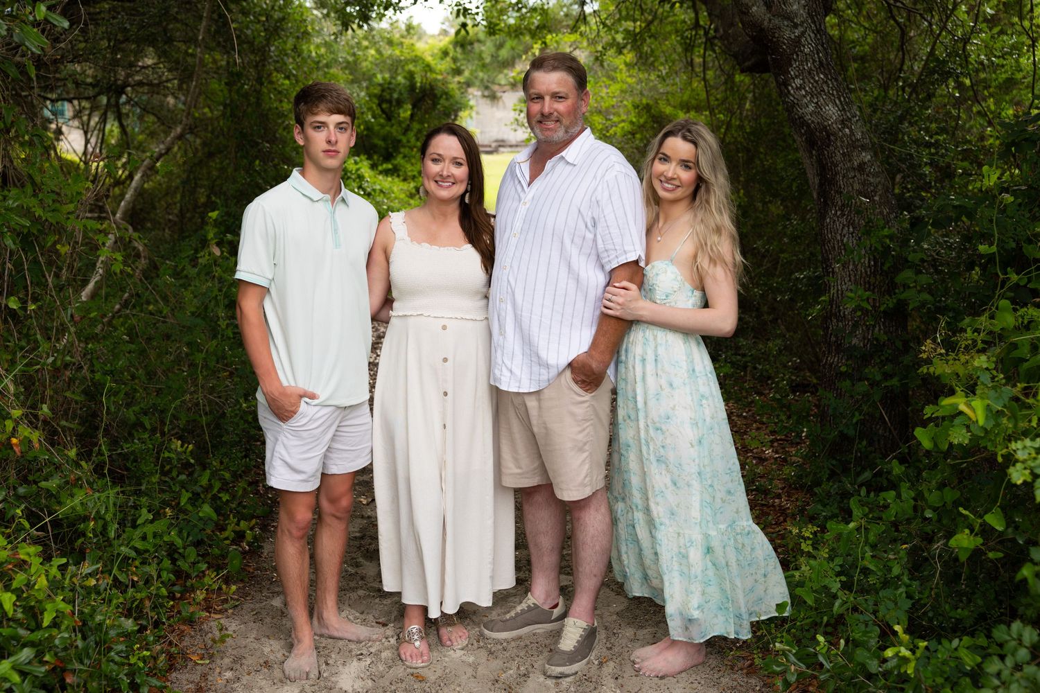 Family of four posing together on a sandy path surrounded by lush green trees in a tropical outdoor setting.