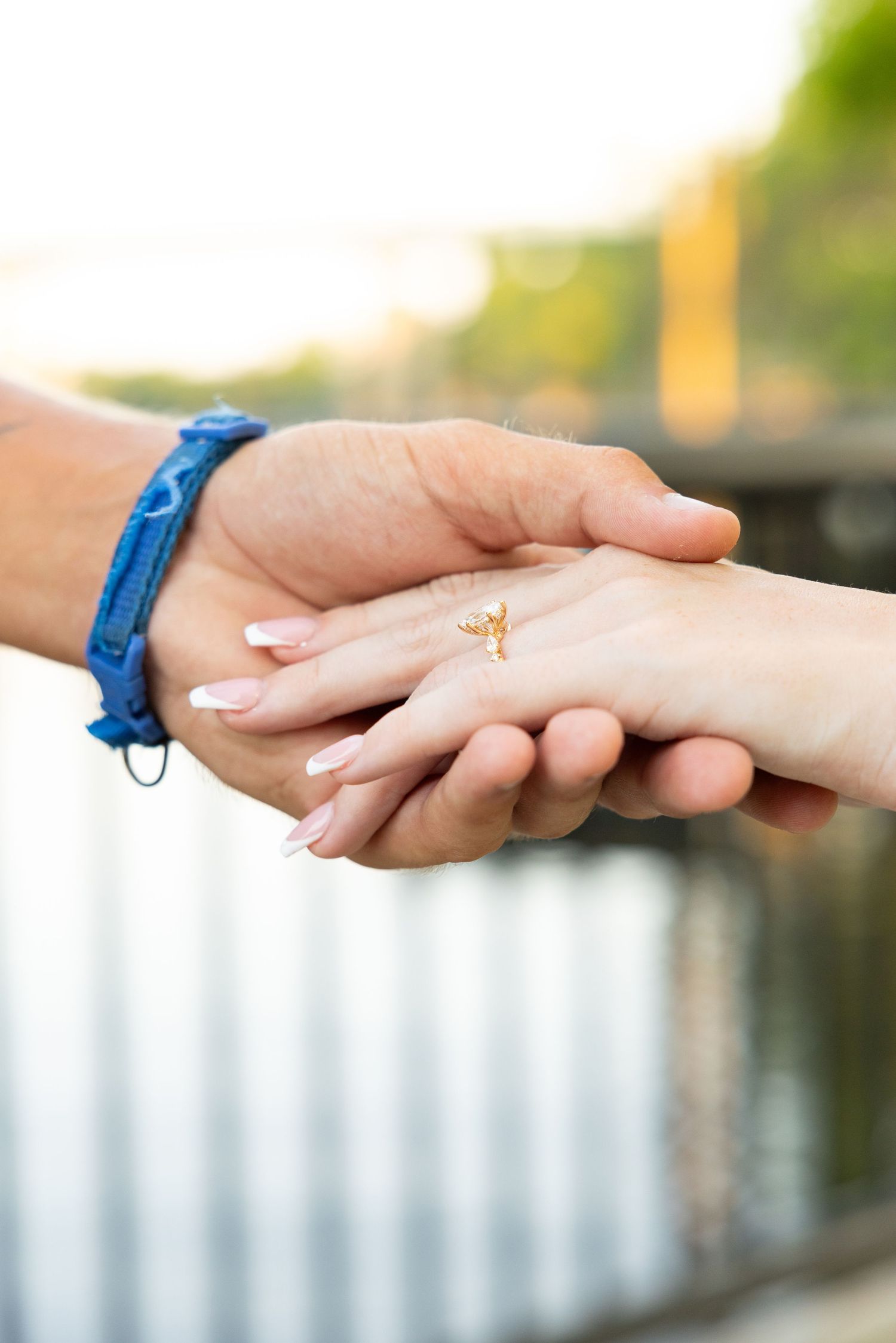 Close-up of two hands holding, woman wearing gold ring with French manicure, man with blue bracelet, outdoor bokeh background.