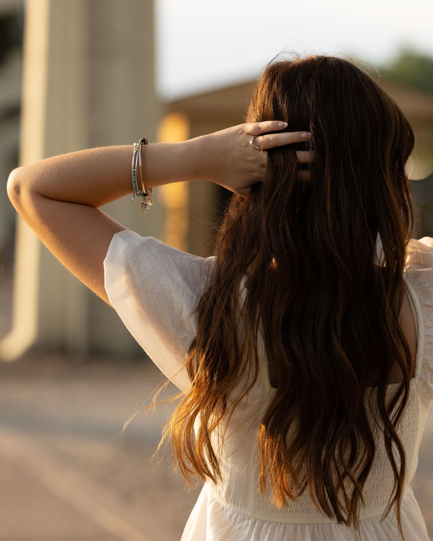 Woman with long wavy brunette hair in white dress, hand raised to head, wearing bracelets, golden hour lighting.