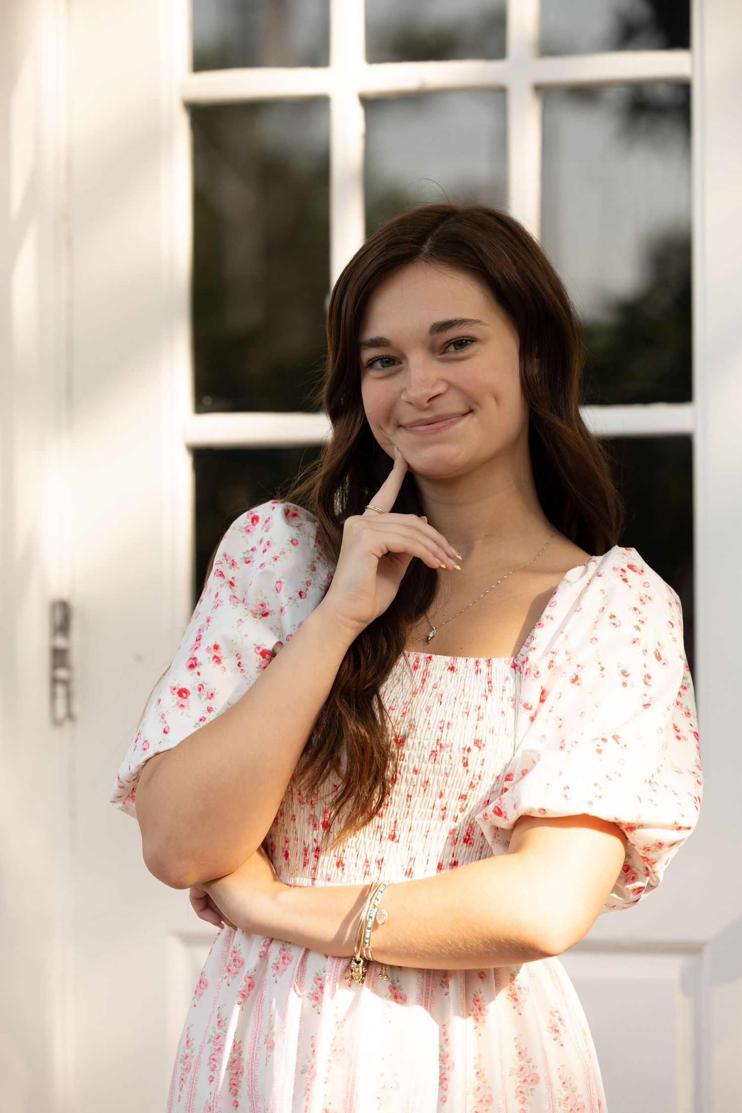 Young woman in white floral dress posing thoughtfully by white French doors outdoors.