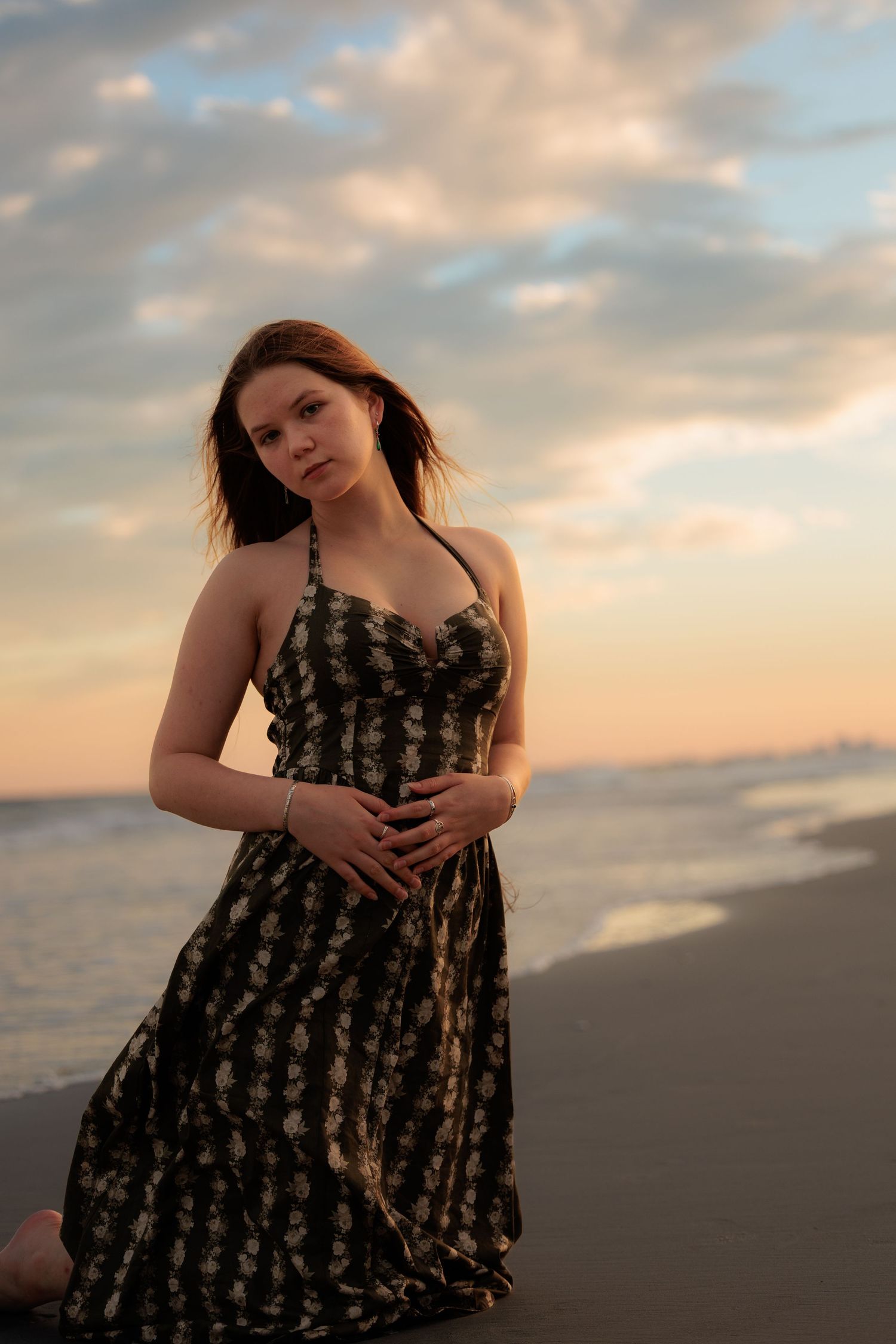 Young woman in dark floral halter dress kneeling on beach at colorful sunset with ocean behind her.