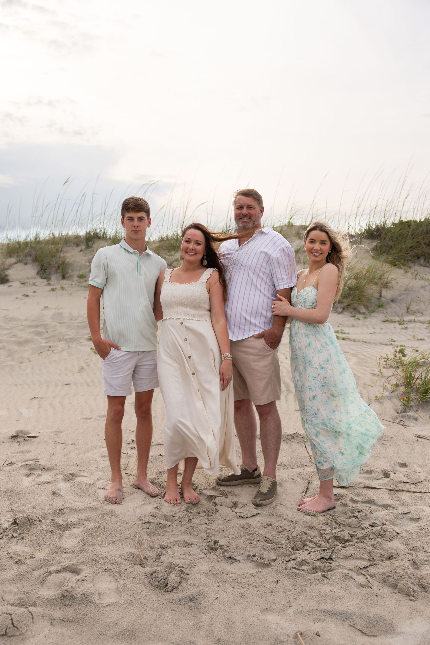 Family of four posing together on a sandy beach at dusk, wearing coordinated pastel outfits with ocean dunes in the background.