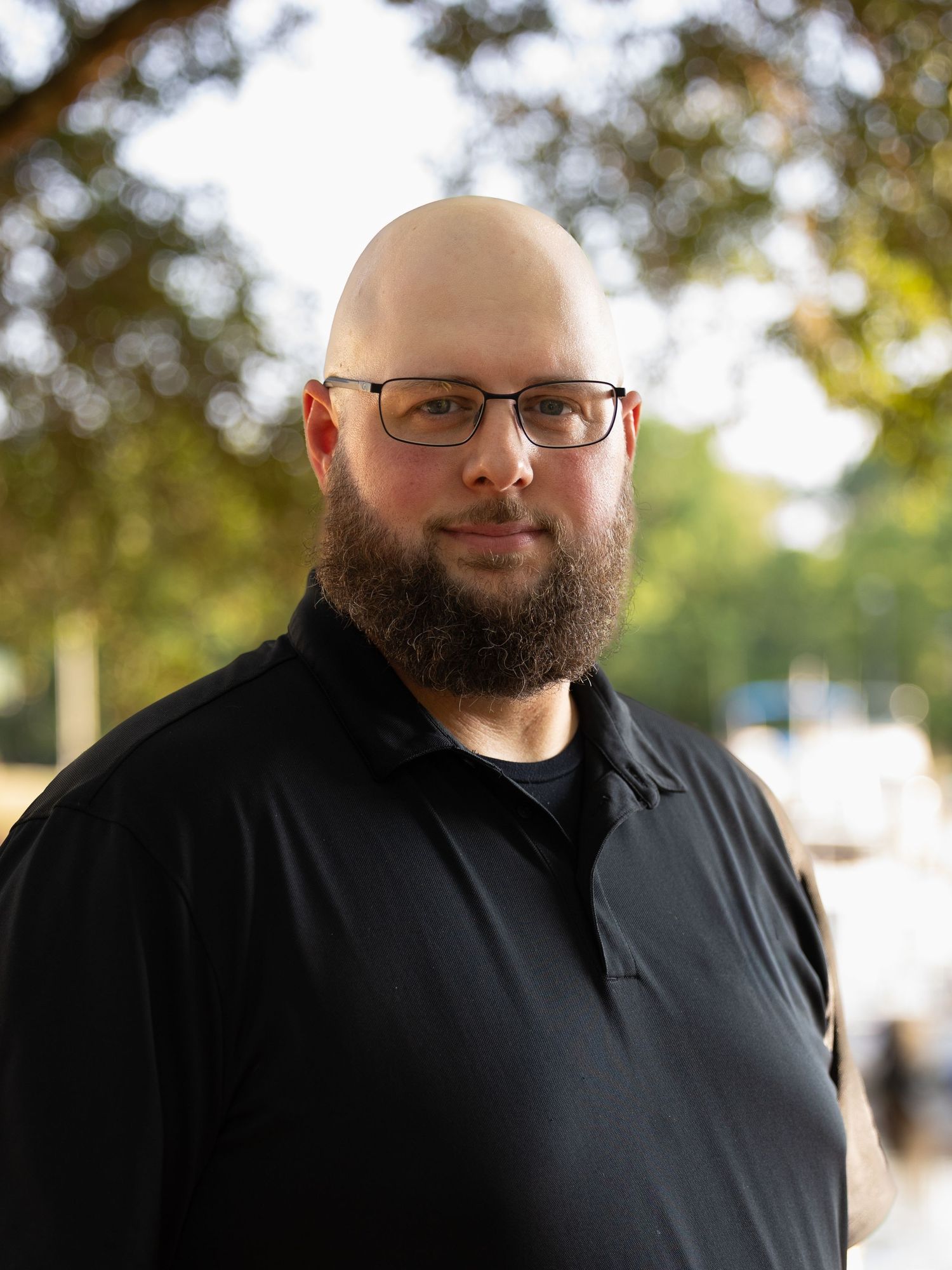 Bald man with beard and glasses wearing a black polo shirt outdoors with blurred trees in background.
