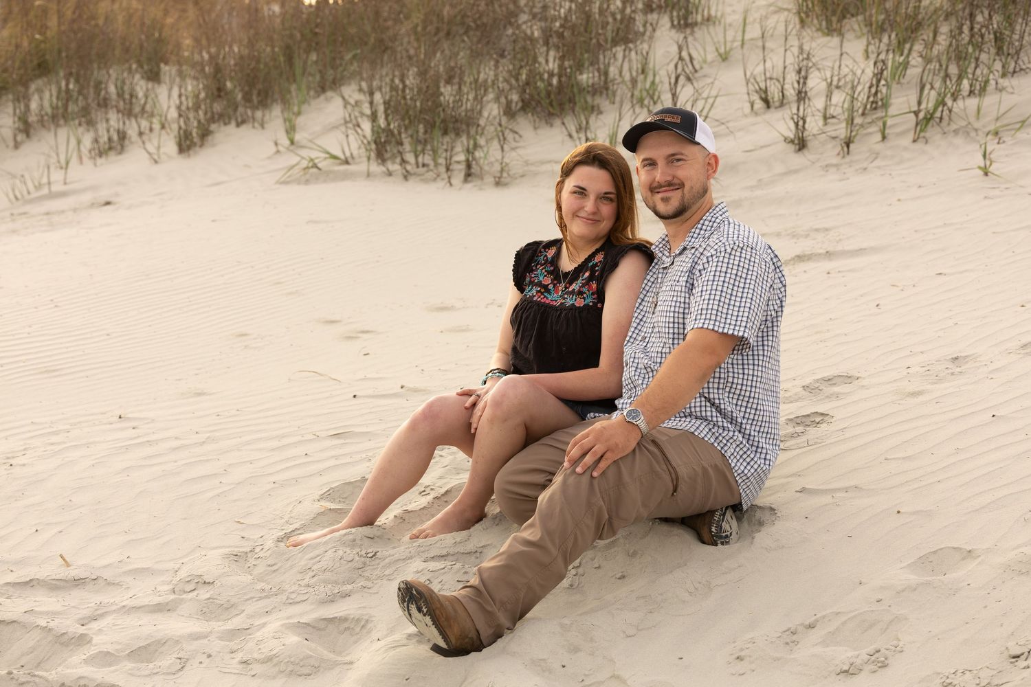 Smiling couple relaxing together on sandy beach dunes, leaning against each other in casual outfits.