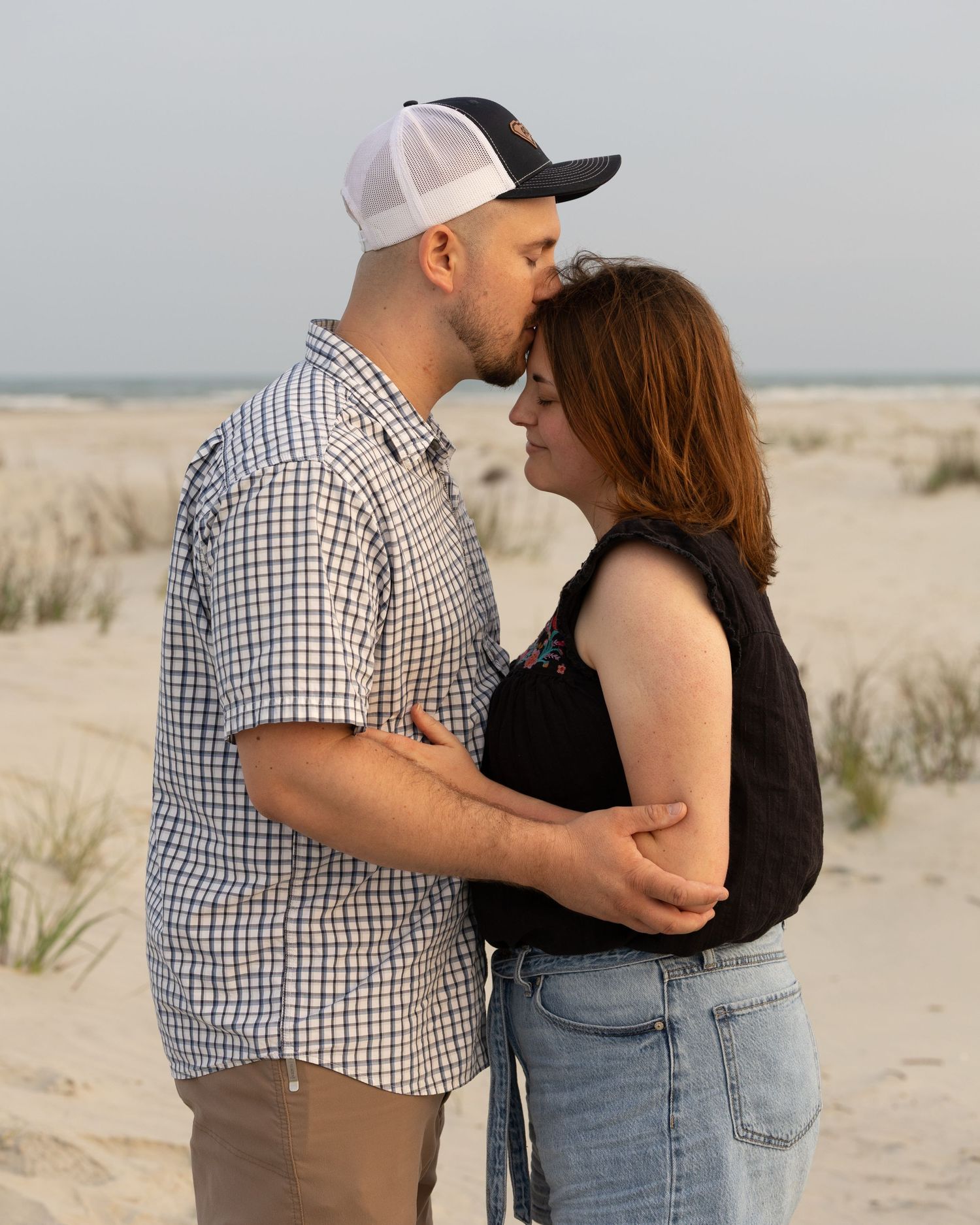 Man in plaid shirt and cap kissing woman on forehead on a sandy beach with dunes in background.