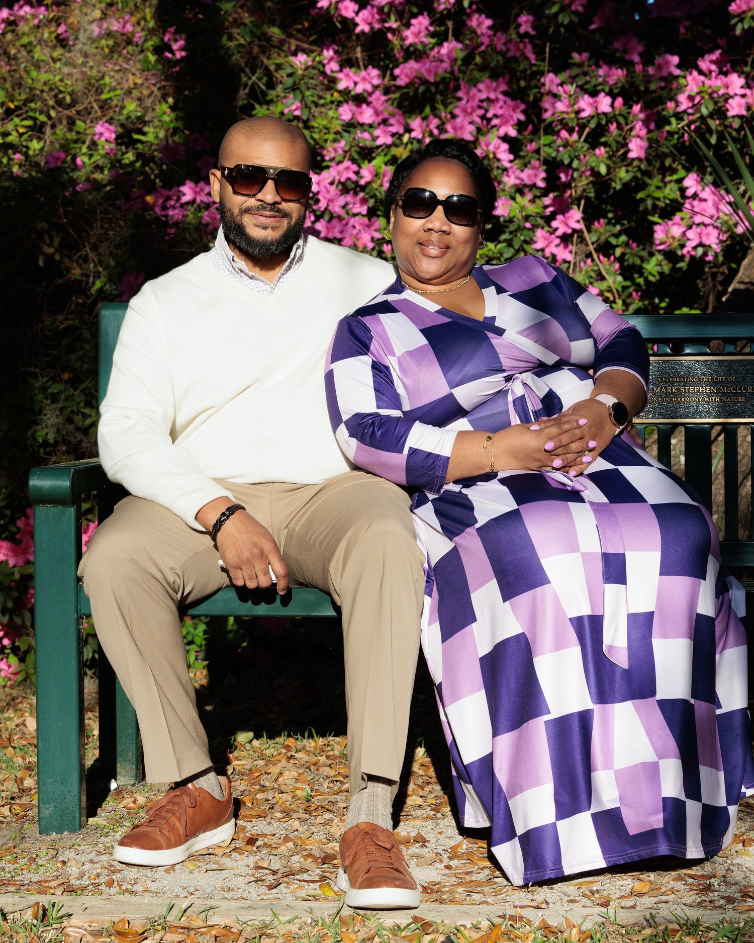 A couple sitting together on a green bench surrounded by pink flowering bushes, dressed in elegant purple and white outfits.