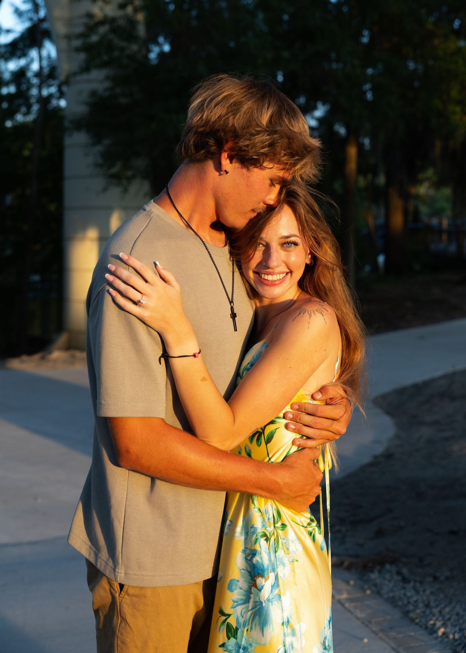 A young couple embraces warmly at golden hour, the smiling woman in a floral dress and man in a gray tee.