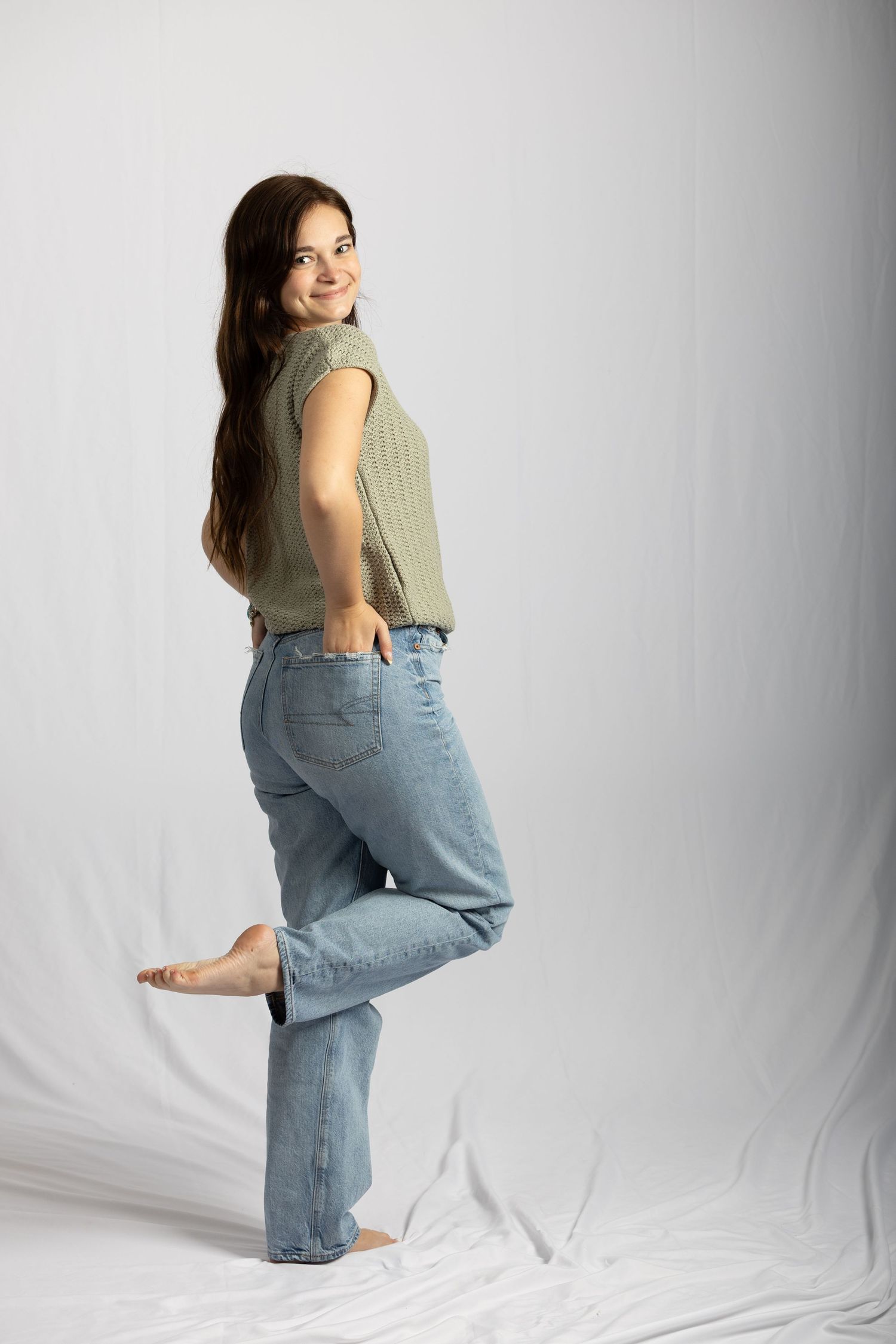 Young woman in green top and blue jeans lifting one foot playfully on white backdrop.