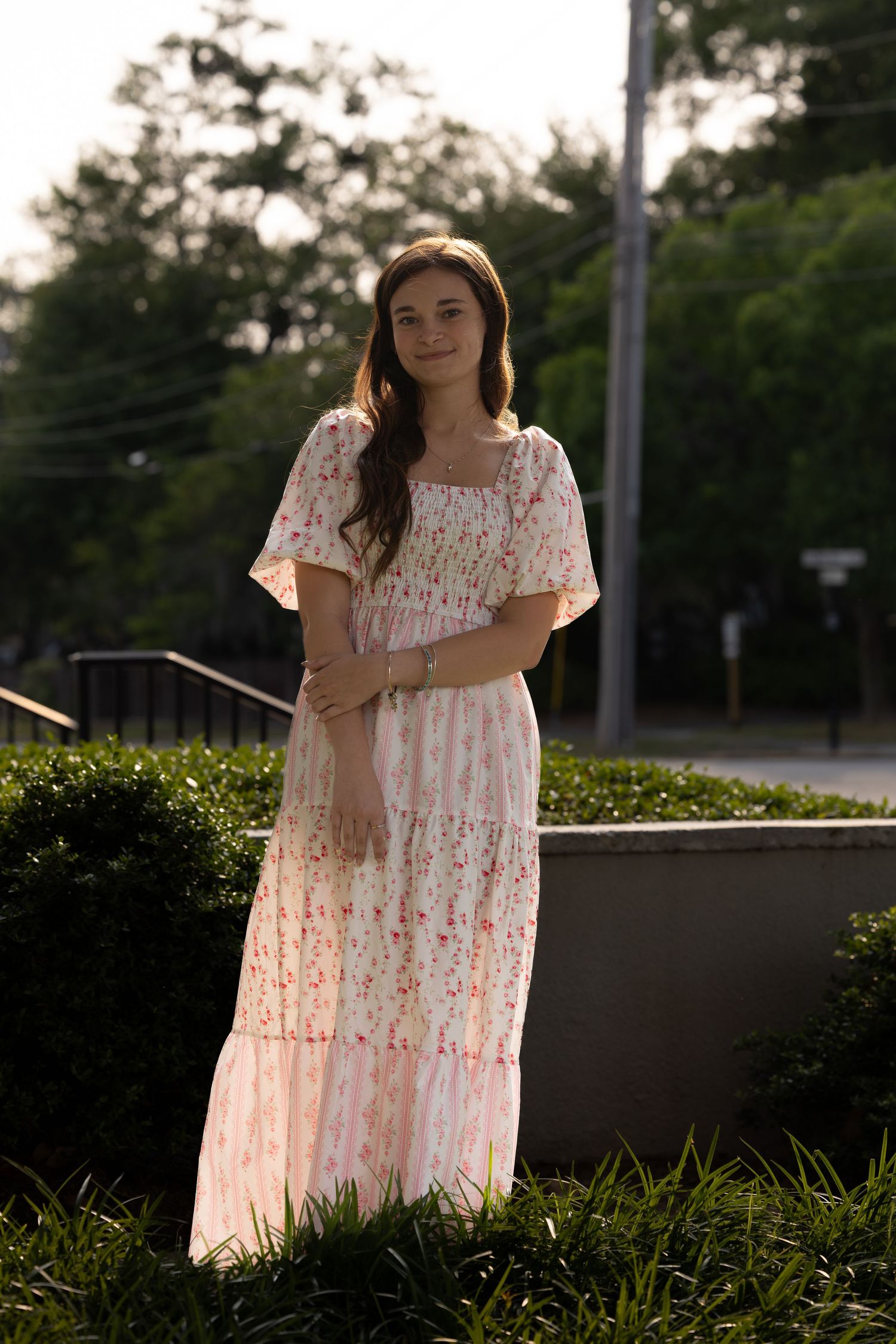 Young woman in white floral maxi dress standing elegantly among green hedges outdoors.