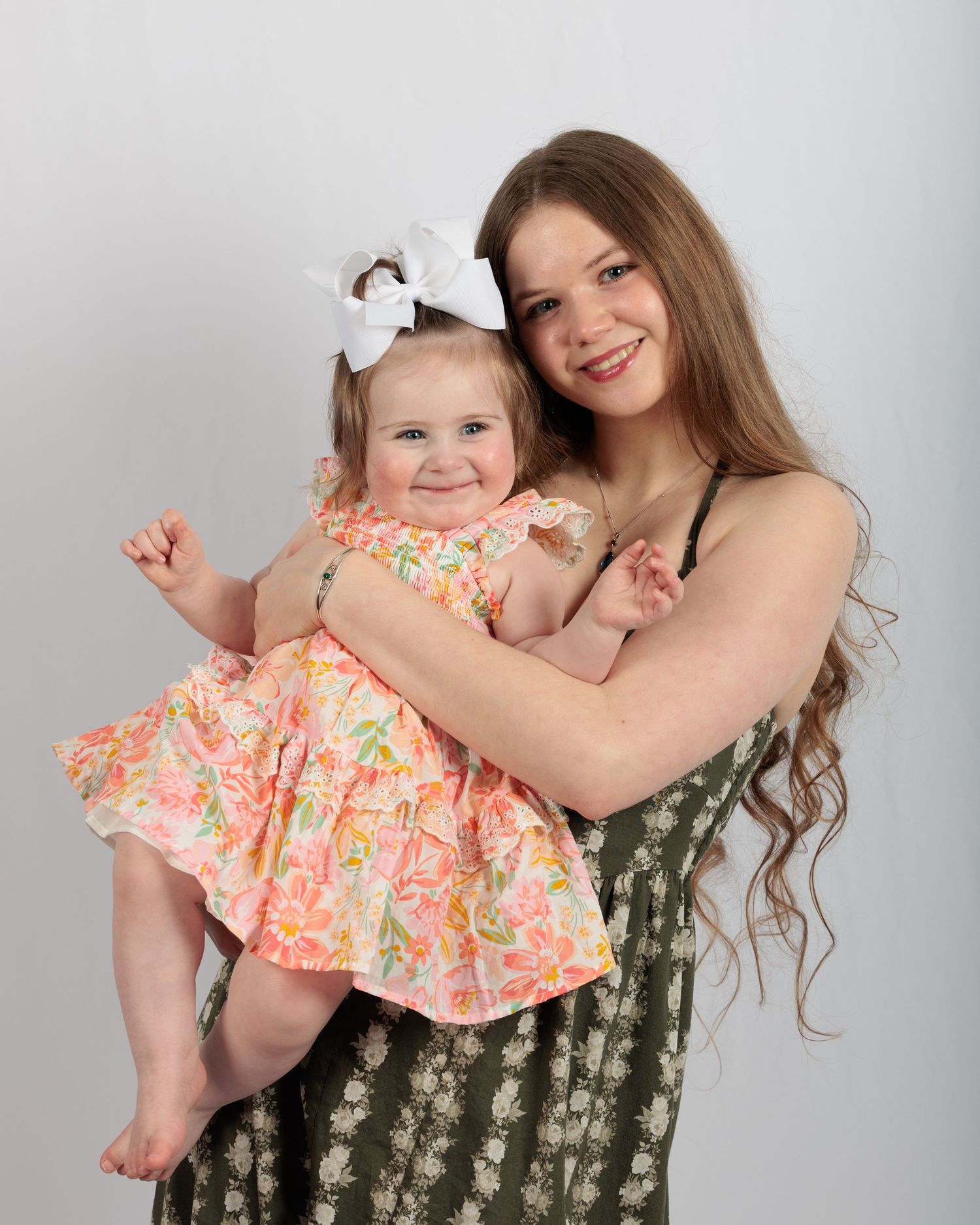 Smiling young woman with long wavy hair holding a happy baby girl in a floral dress with white bow.