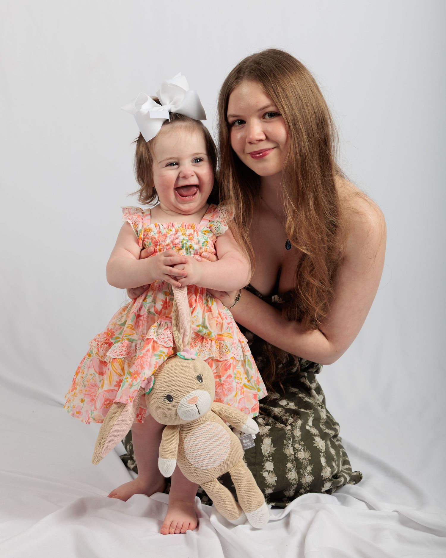 Young woman and laughing toddler girl in floral dress with bow and bunny stuffed animal pose for portrait.