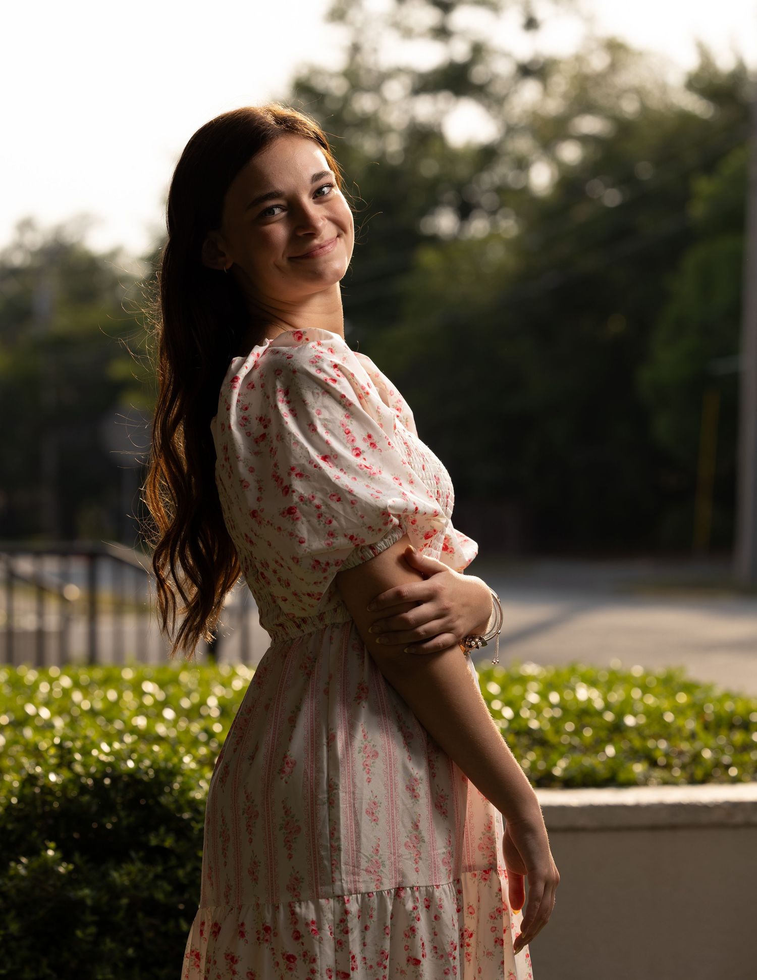 Young woman in white floral dress smiling over her shoulder in warm evening light.