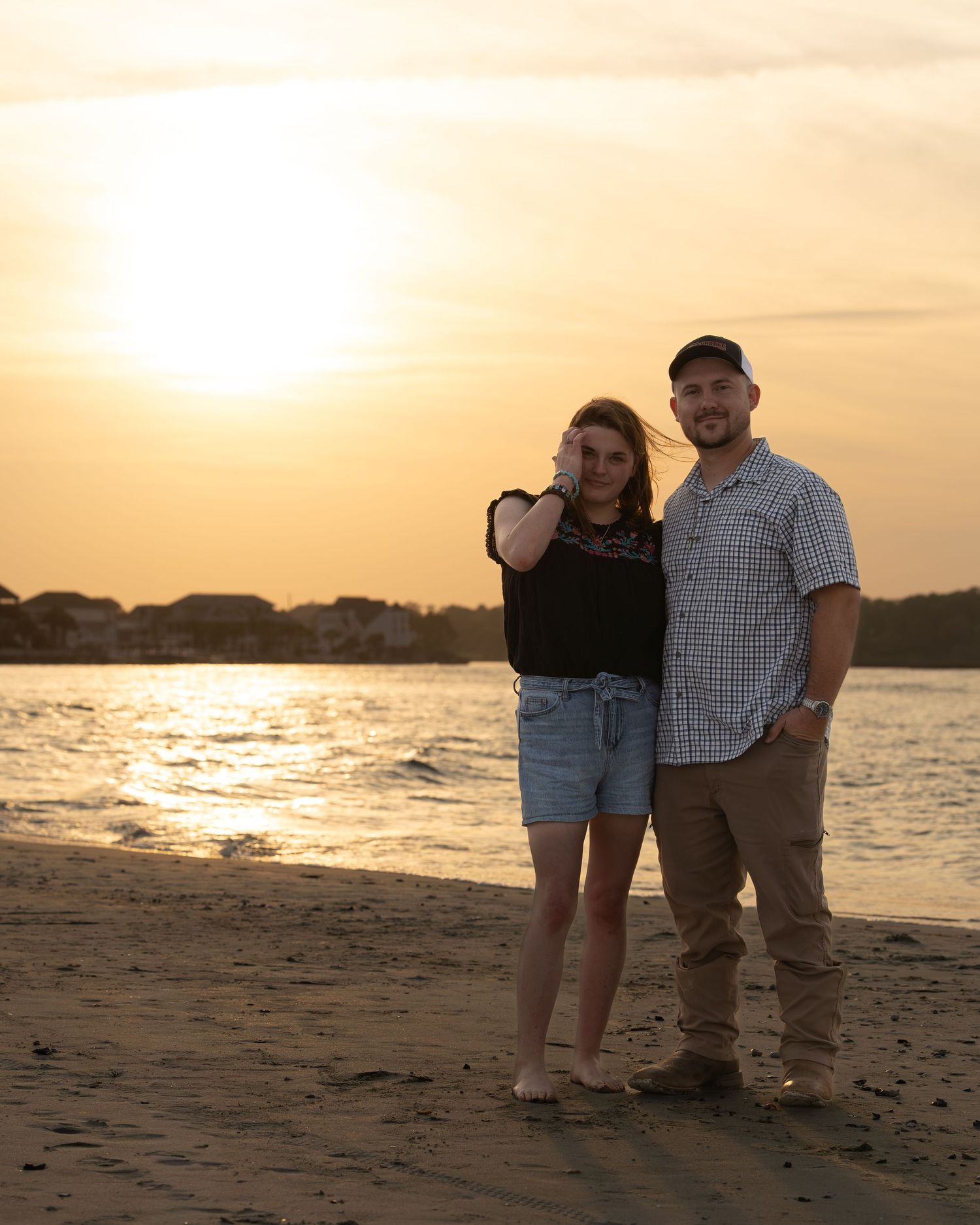Couple posing together on a sandy beach at golden sunset with calm water reflecting warm orange light.