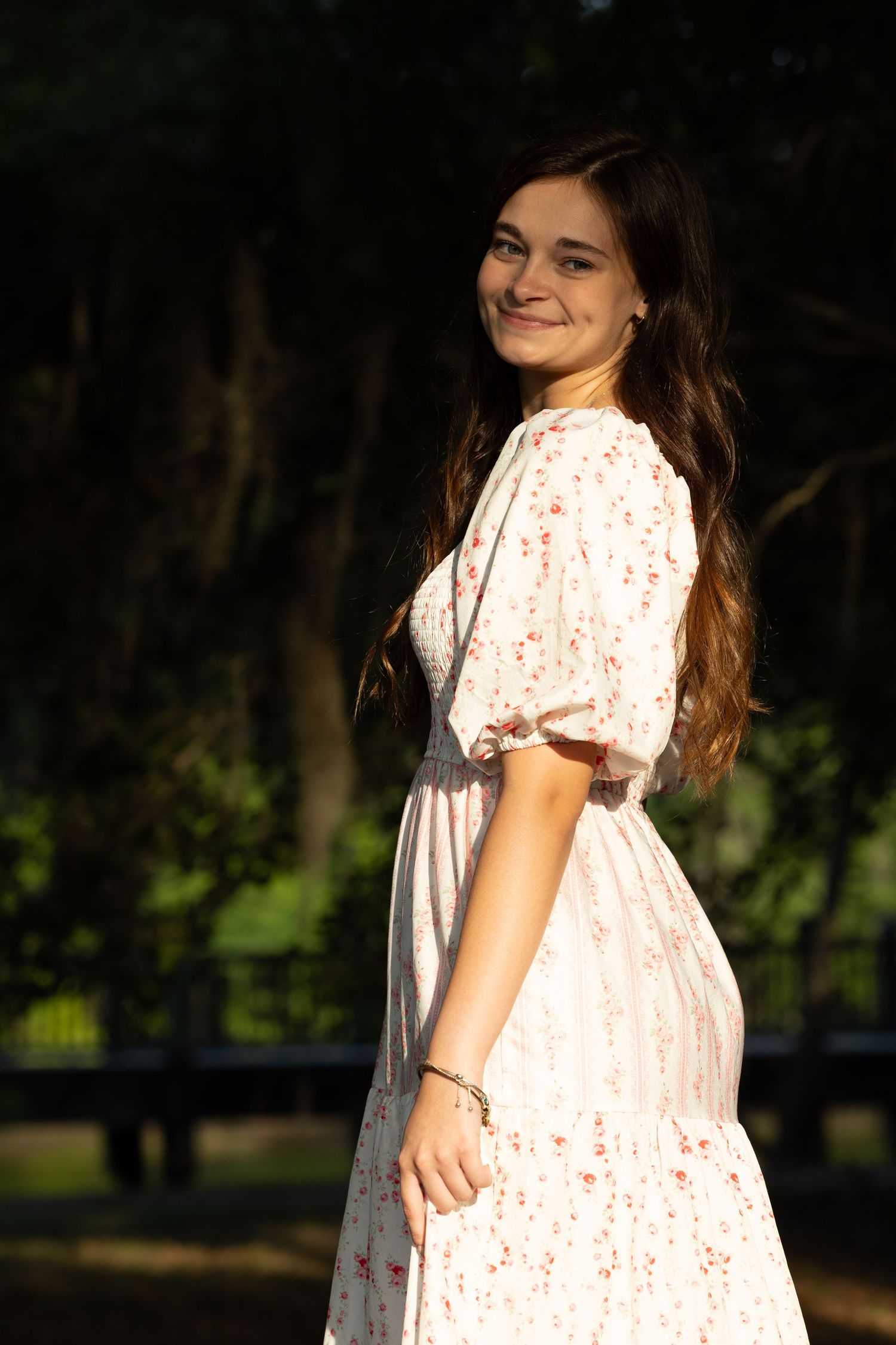 Young woman in white floral midi dress smiling in golden hour sunlight in park.