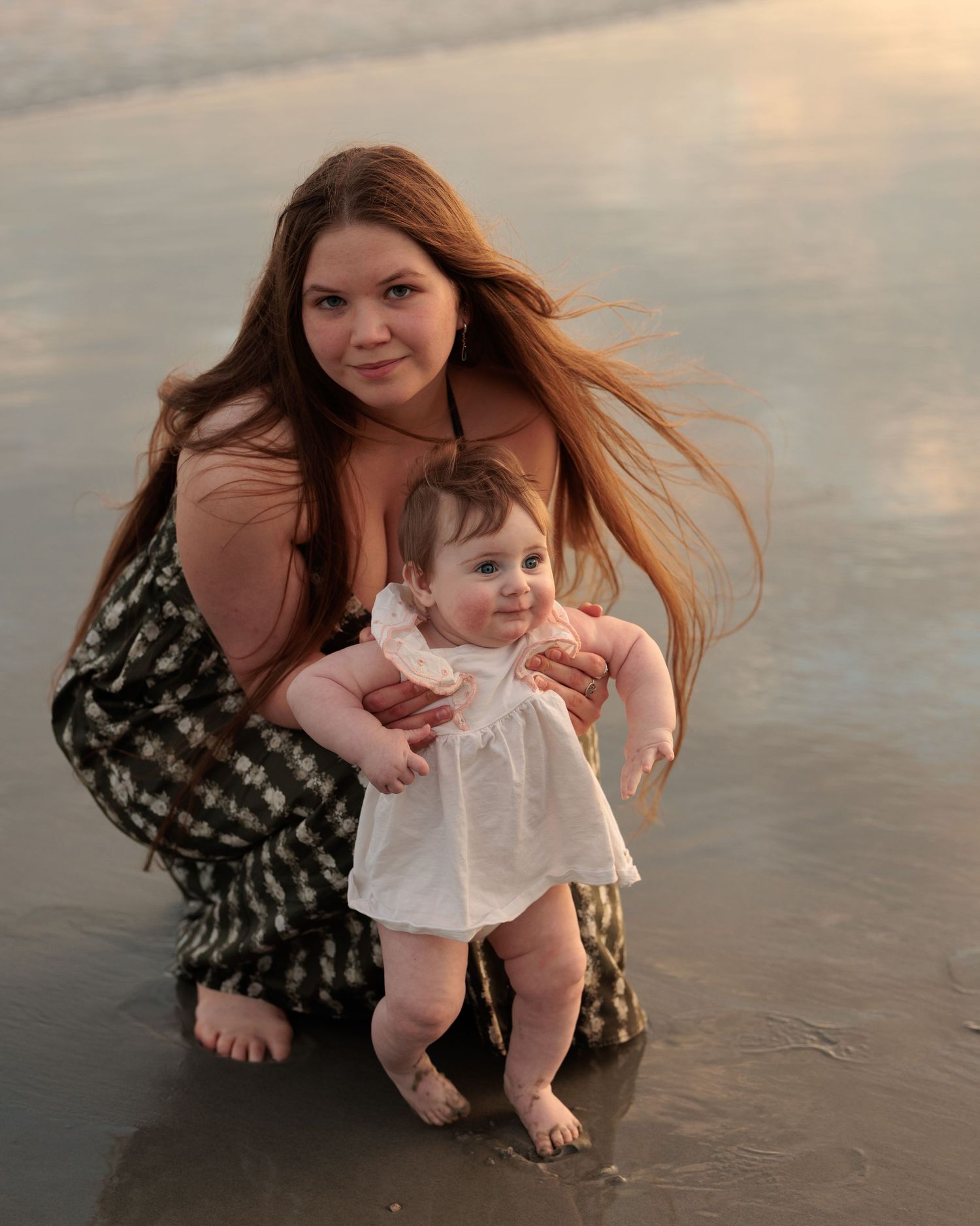 Young mother with long red hair crouching on wet beach at sunset, holding smiling baby in white dress.