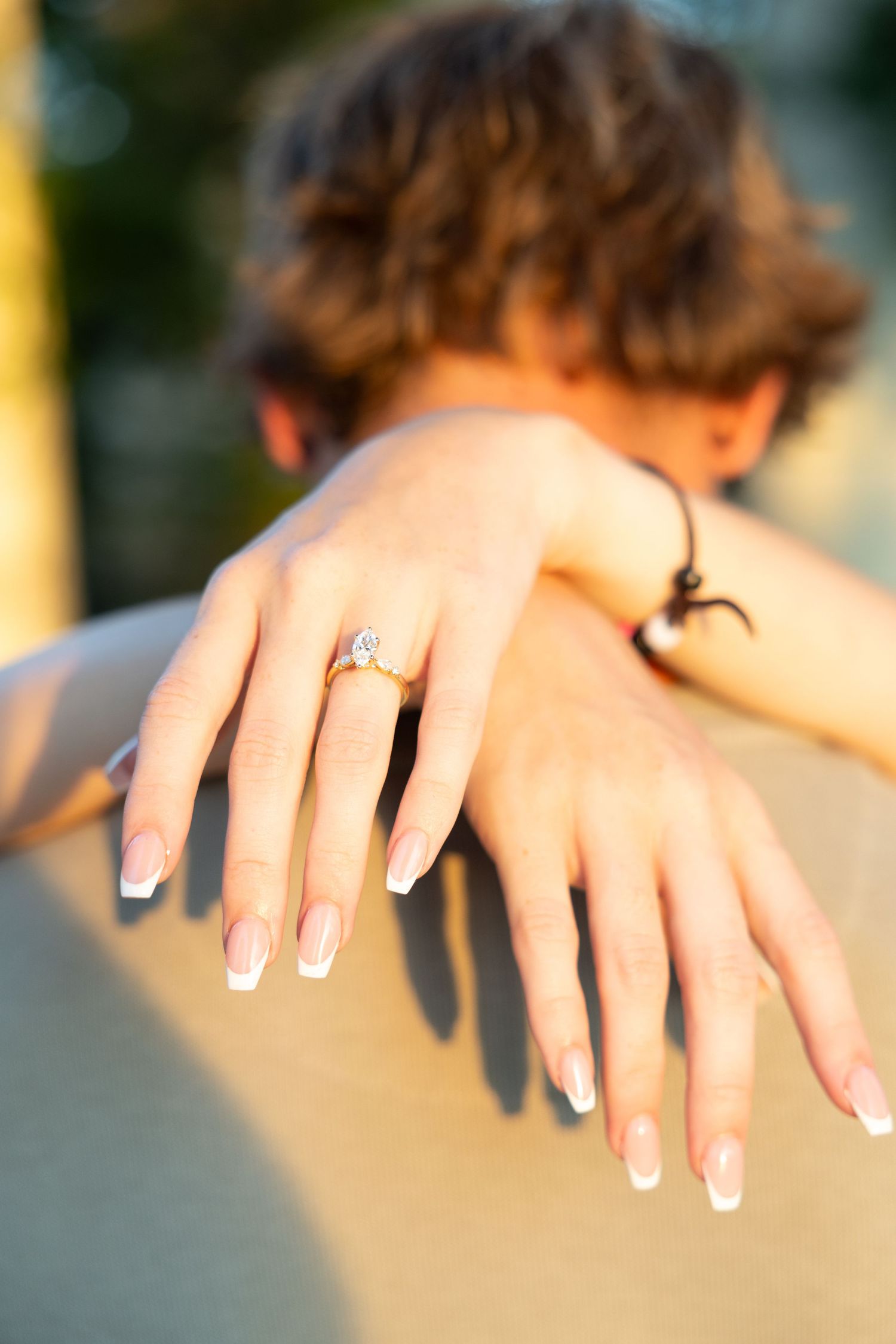 Close-up of a woman's hands with French manicure displaying an engagement ring while embracing her partner outdoors.