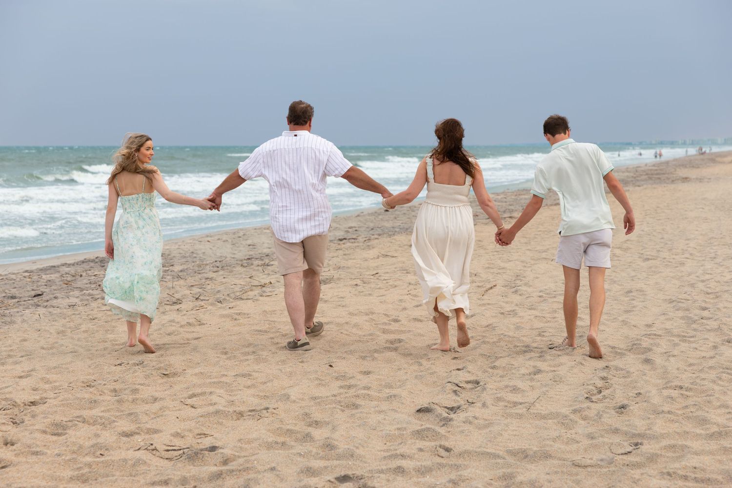 Four people holding hands walk along a sandy beach toward ocean waves on a cloudy day.
