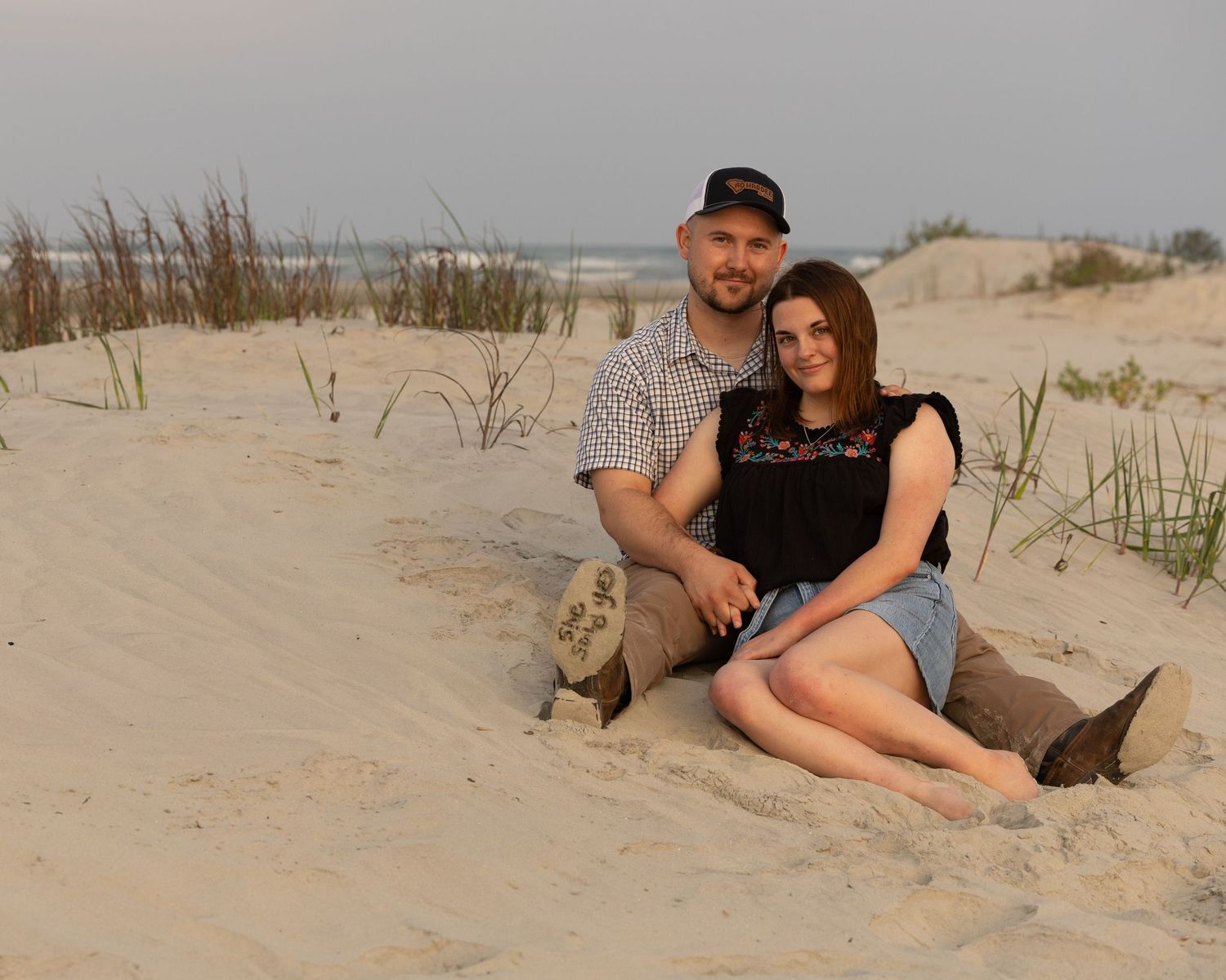 Couple sitting together on sandy beach dunes at dusk, smiling warmly in a romantic outdoor portrait.