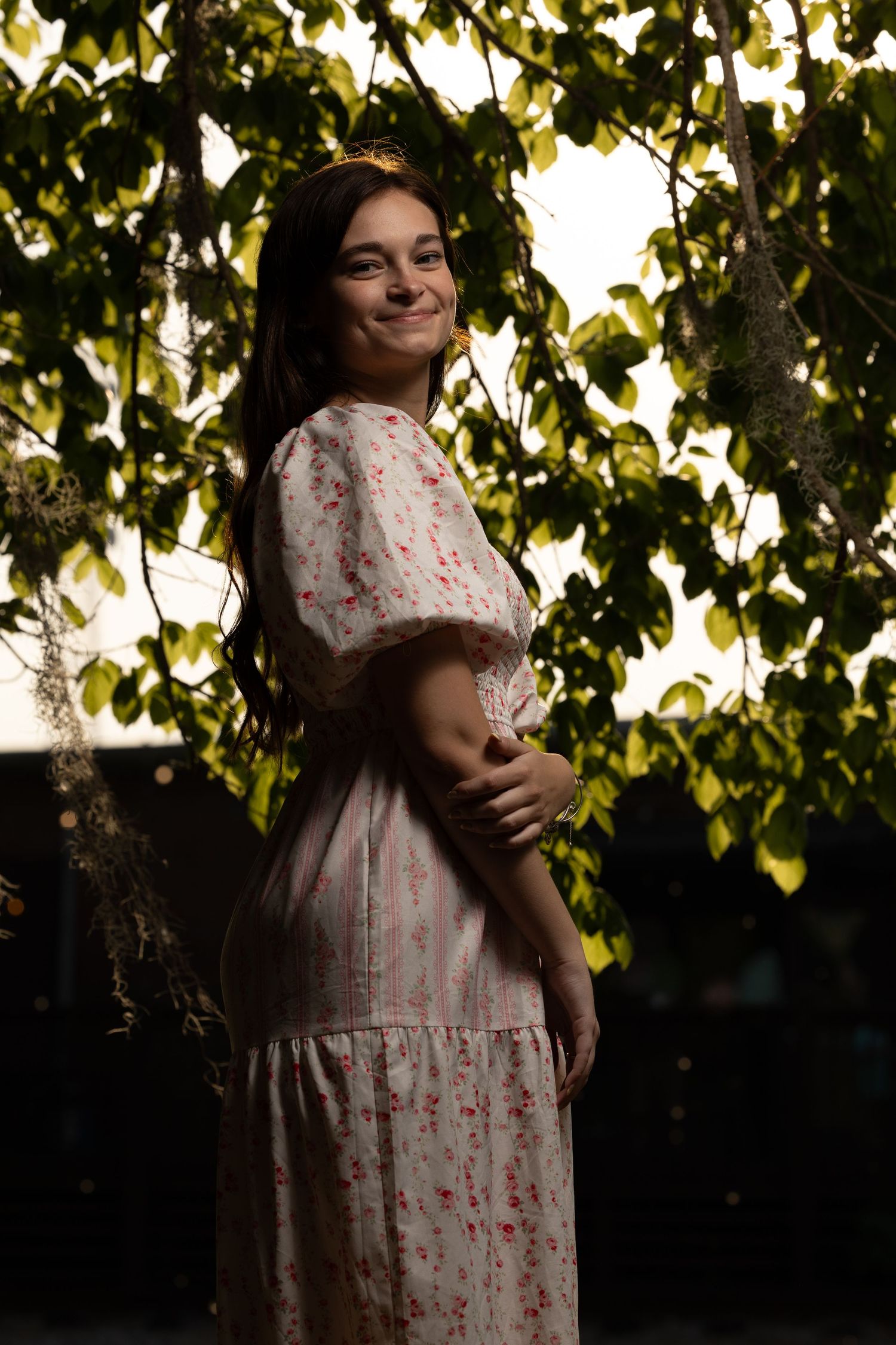 Smiling young woman in floral dress standing among green hanging leaves outdoors.
