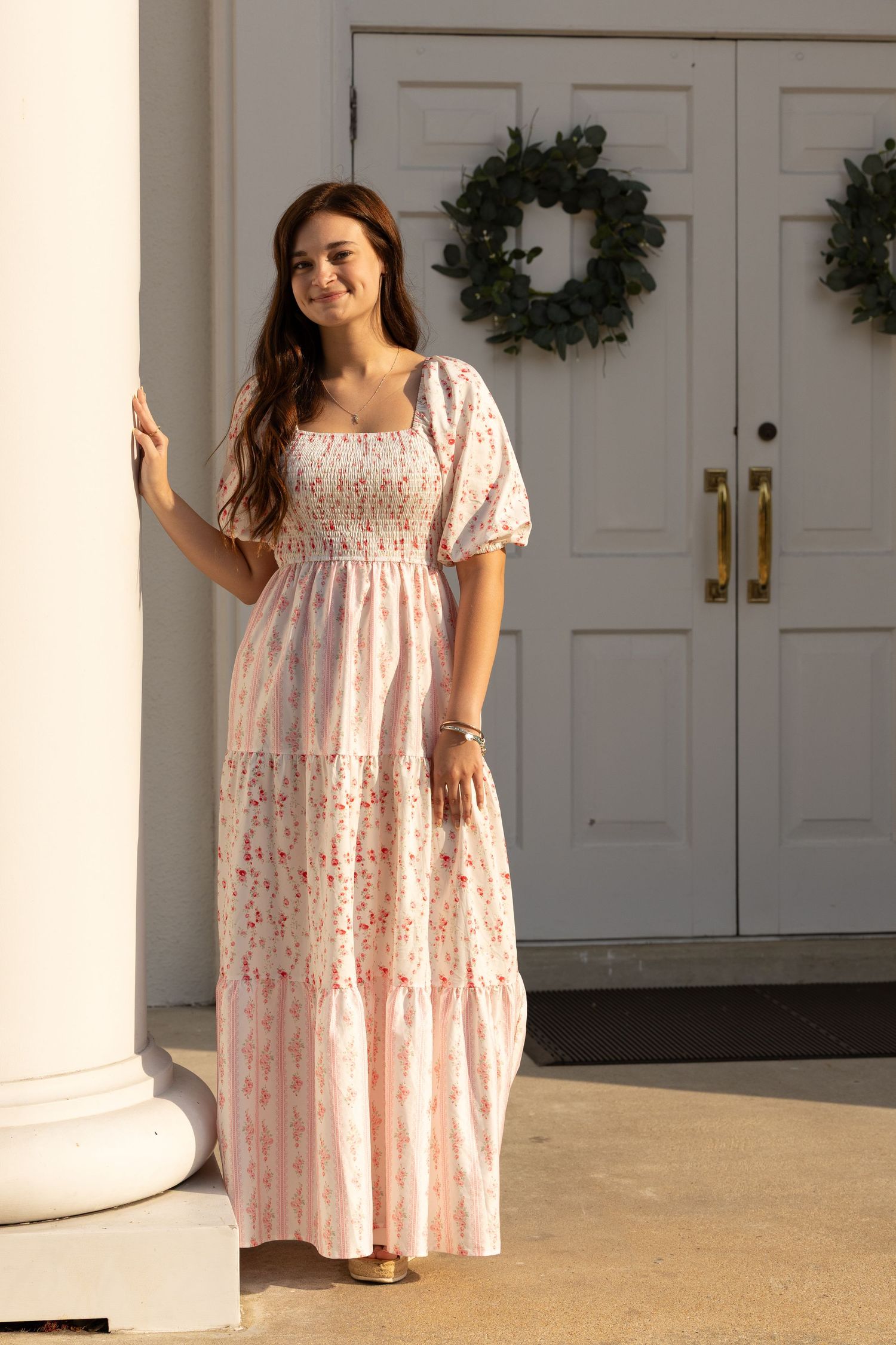 Young woman in pink floral maxi dress posing by white columns and door with wreath.