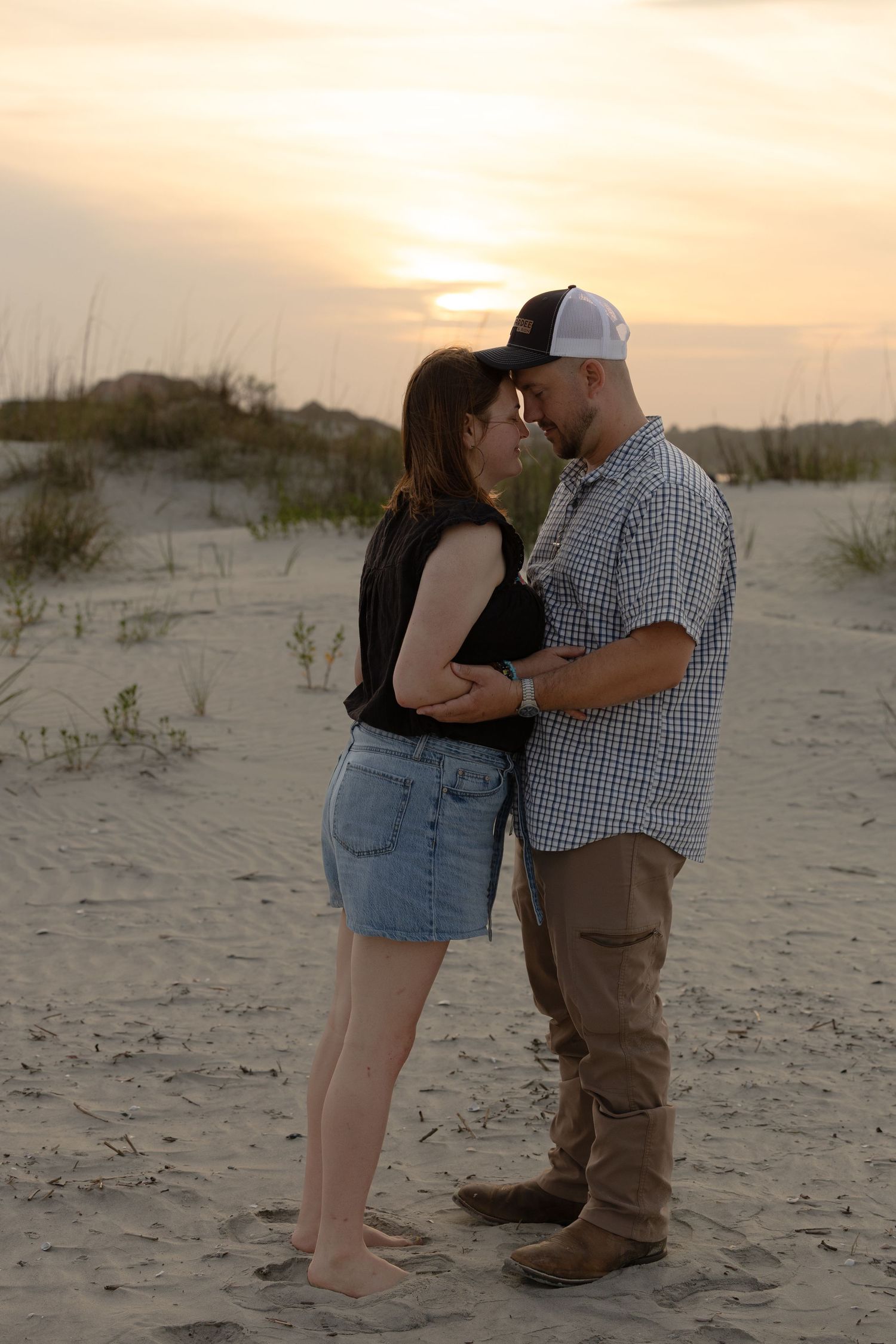 Romantic couple embracing forehead-to-forehead on sandy beach at golden sunset with dunes behind them.