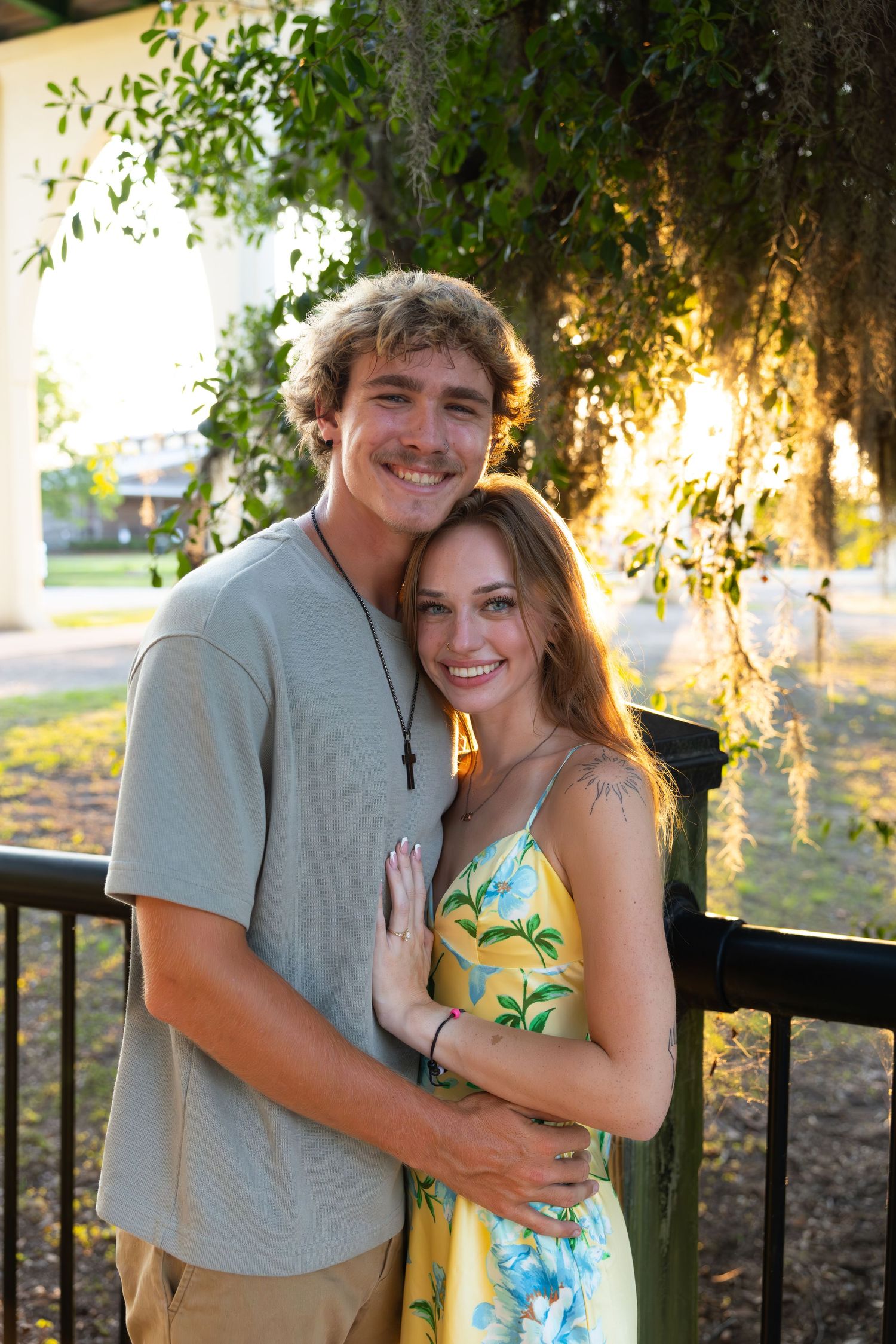 A smiling young couple embraces outdoors at golden hour, woman in floral yellow dress, man in gray tee with cross necklace.