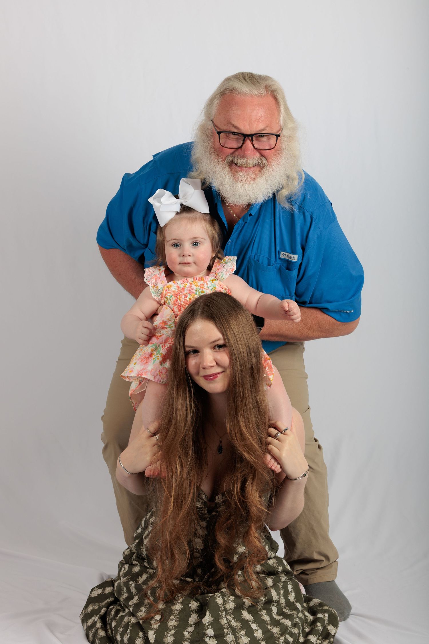 Three generation family portrait with elderly bearded grandfather, young woman, and toddler girl in floral dress.