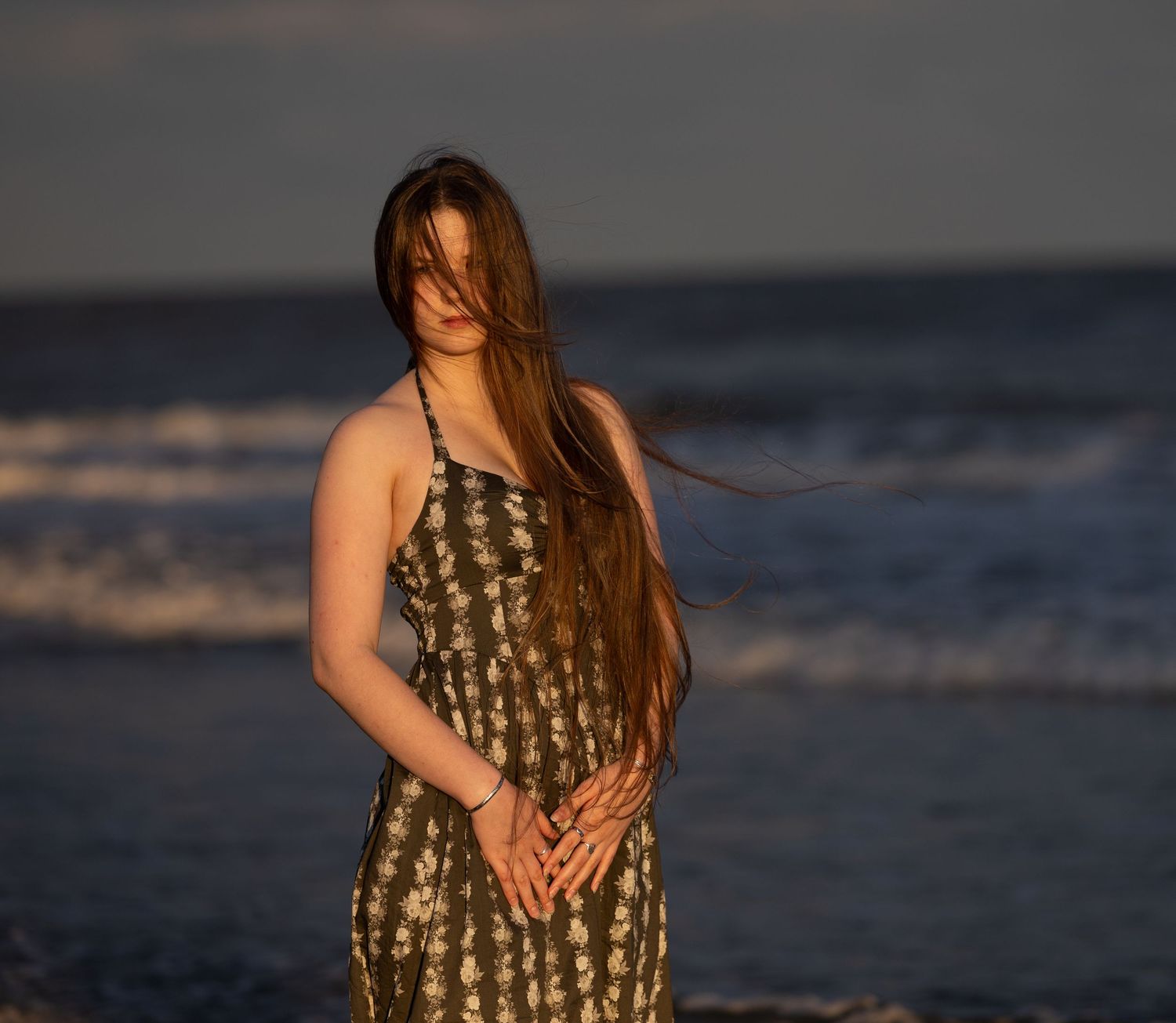 Young woman in floral sundress standing on beach at sunset with hair blowing in the wind.