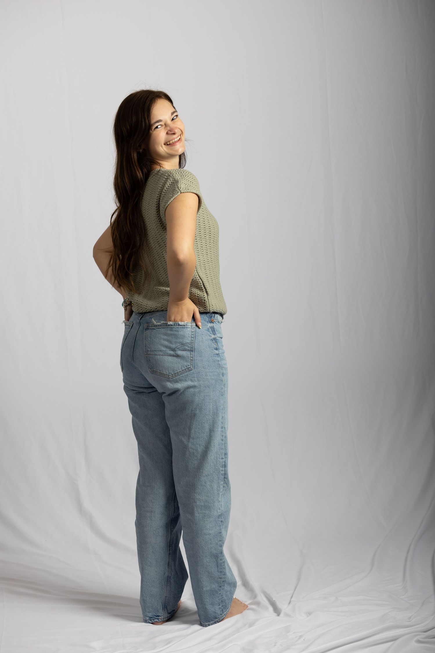 Young woman in sage green top and light blue jeans posing sideways on white backdrop.