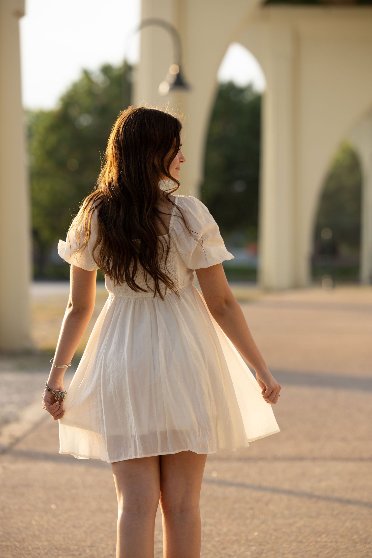 Young woman in white dress walking away from camera near arched columns at sunset.