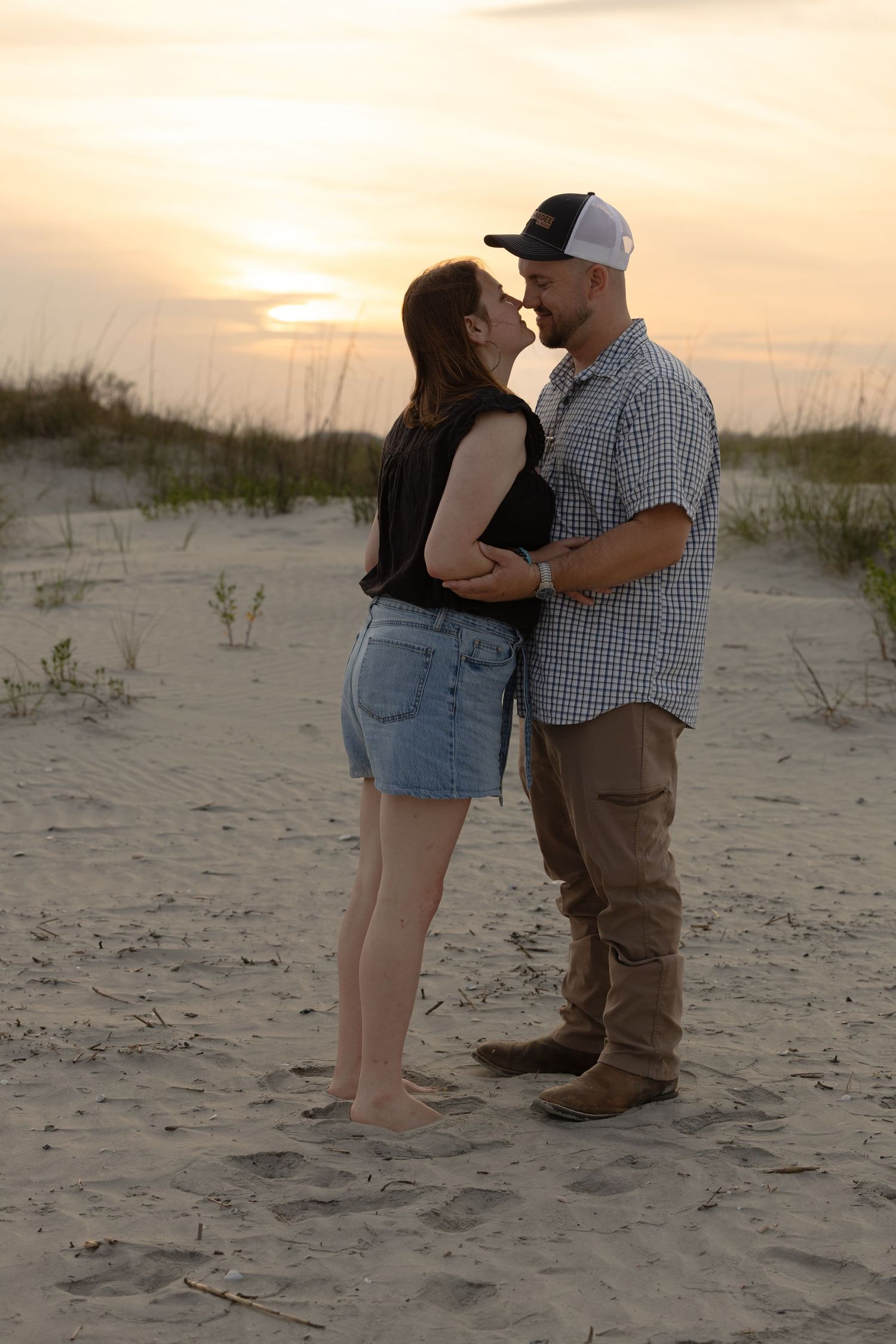Couple sharing a tender moment on sandy beach dunes at sunset, standing close and almost kissing.