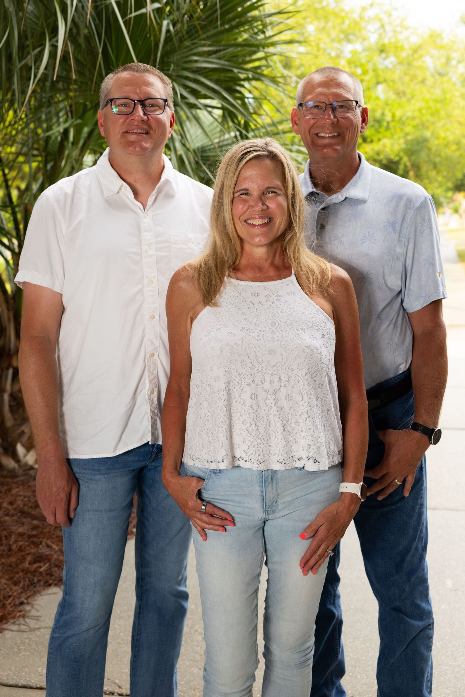 Three smiling adults pose outdoors in casual white and blue outfits surrounded by lush green trees on a sunny day.