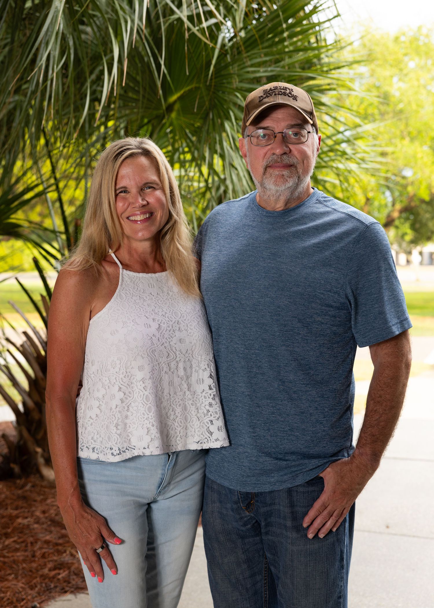 A smiling couple poses outdoors near a palm tree, woman in white lace top and jeans, man in blue t-shirt and cap.