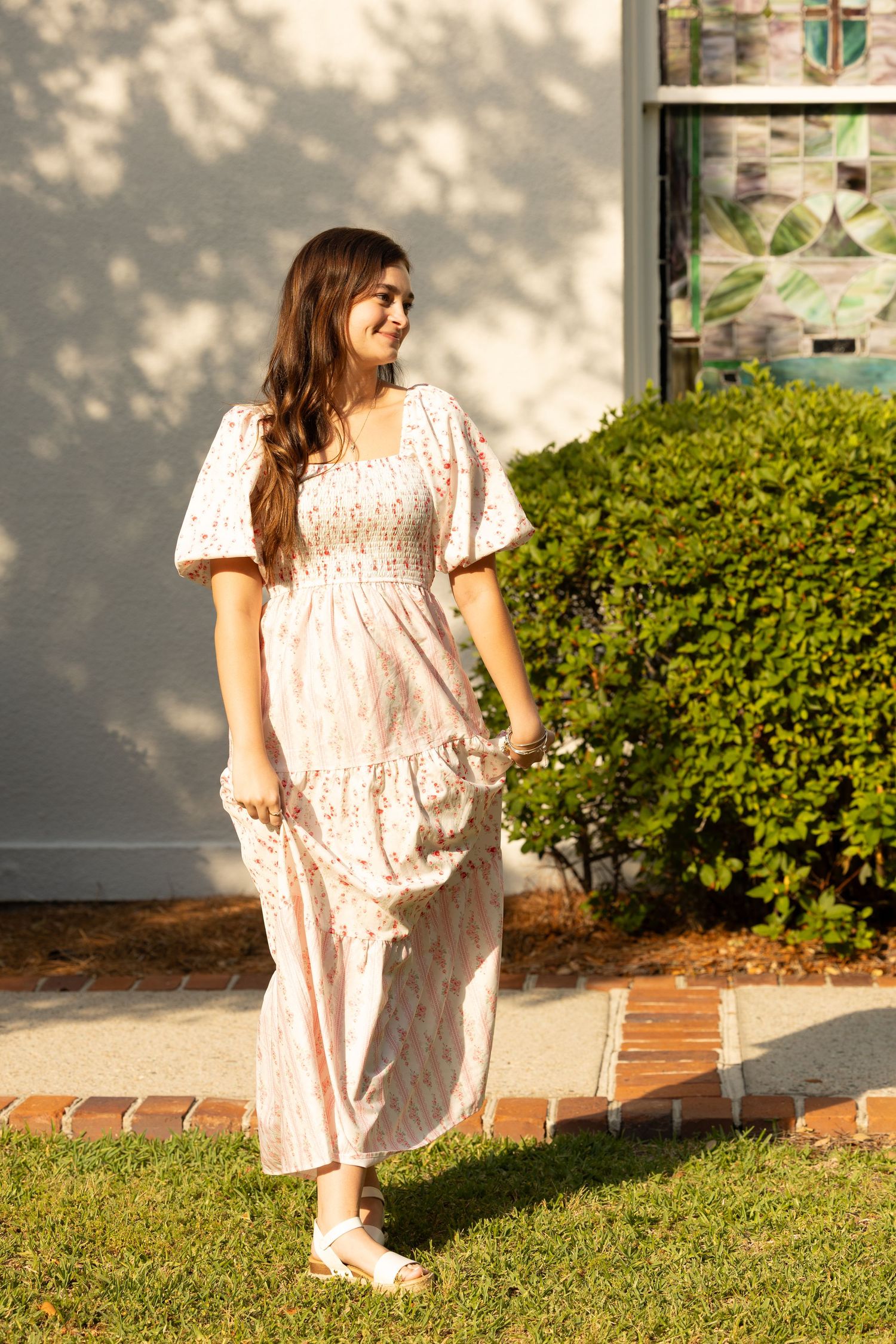 Young woman in white floral maxi dress walking gracefully on green lawn near shrubs.