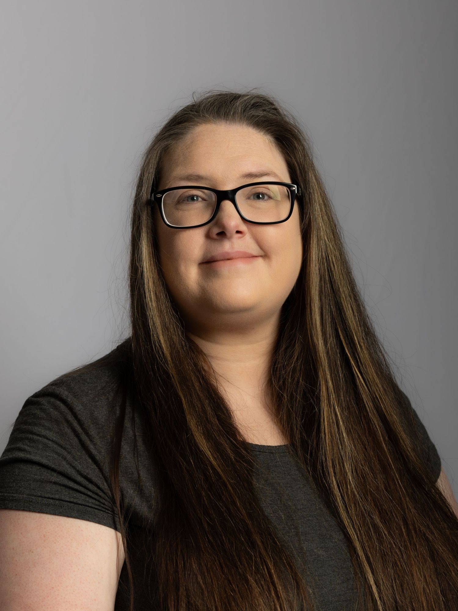 Woman with long brown hair and black glasses wearing a dark gray top poses for a professional headshot photo.
