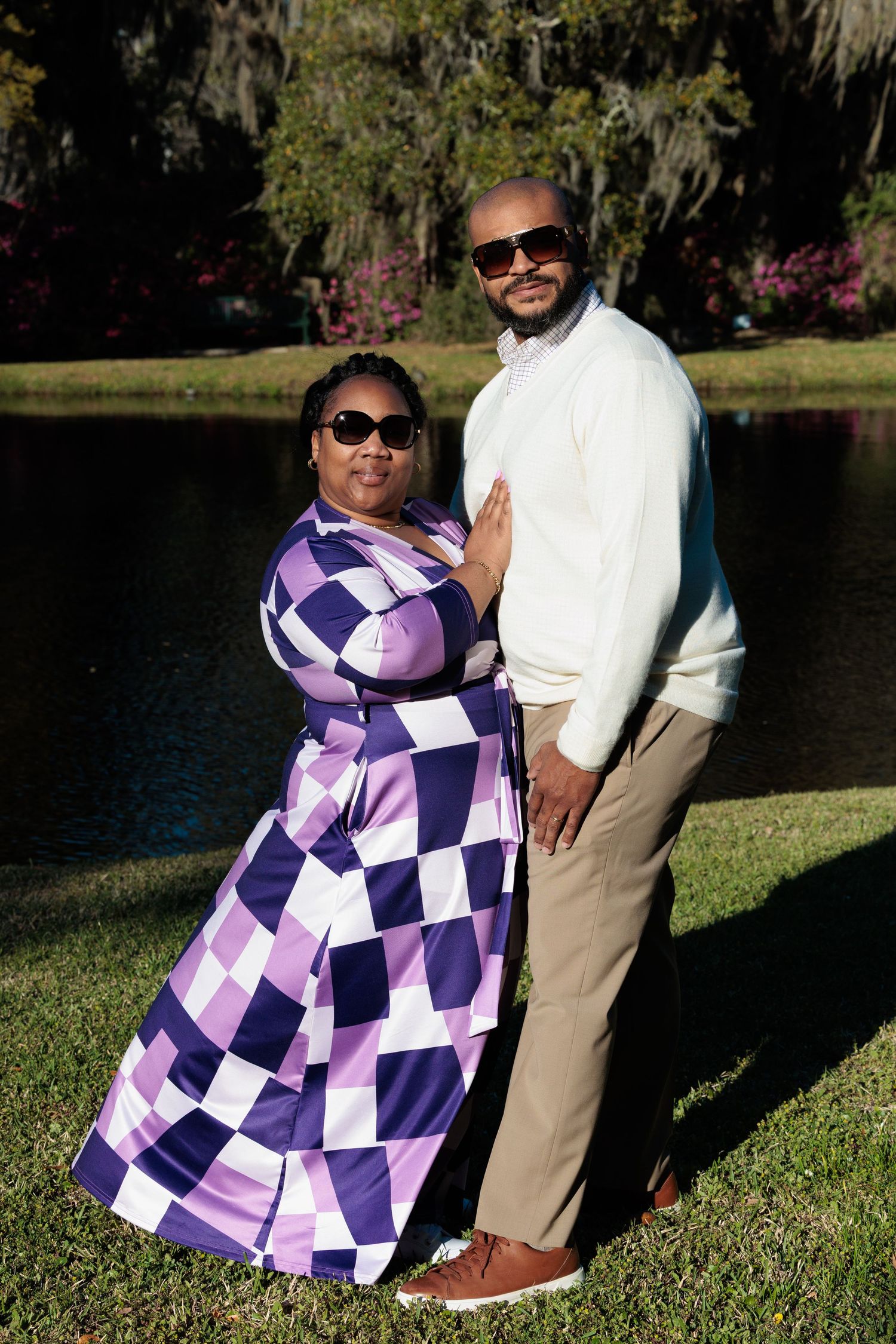 Couple in coordinating purple checkered dress and white sweater pose outdoors near pond in park.