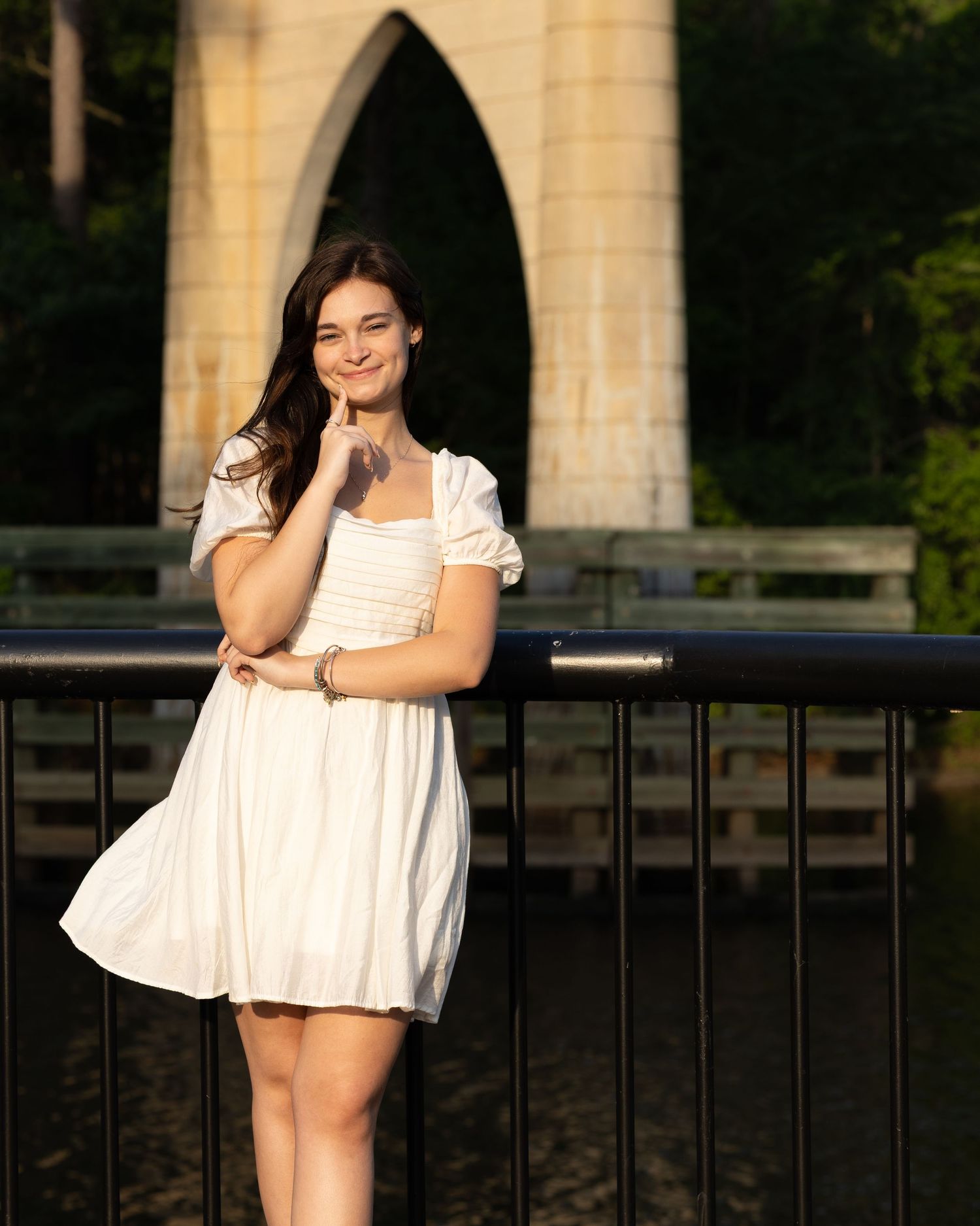 Smiling young woman in white mini dress posing by railing with arched bridge backdrop.