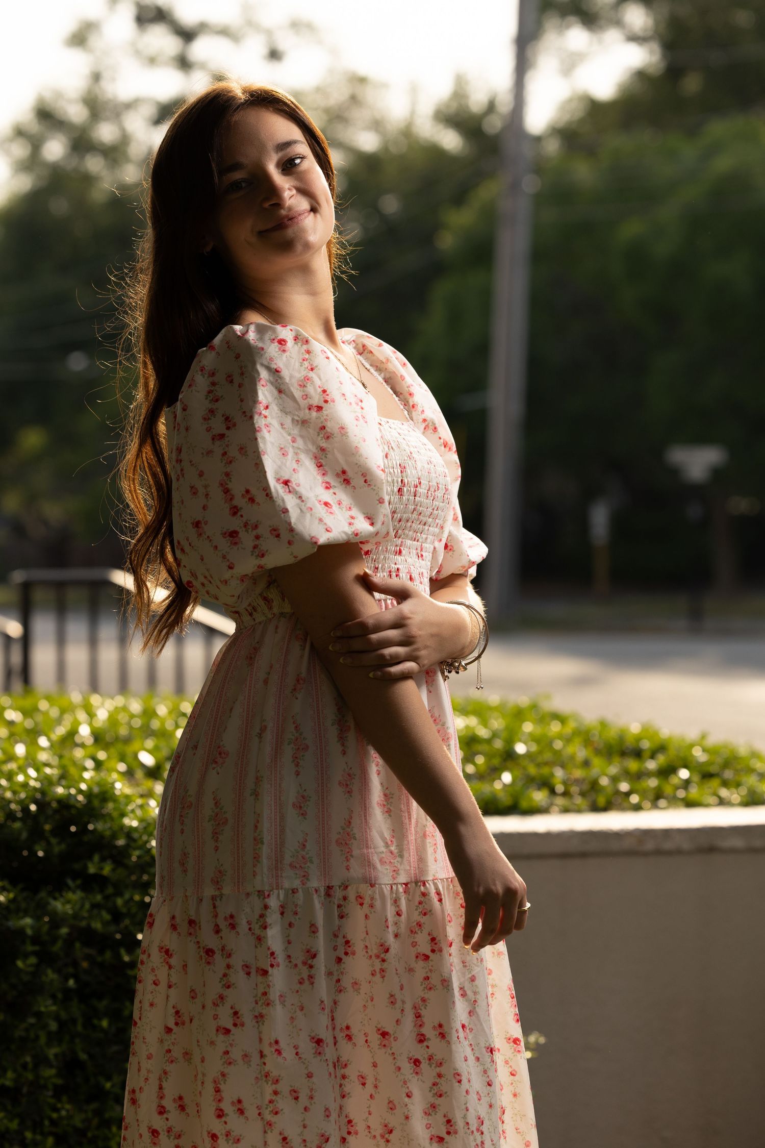 Smiling young woman in pink floral dress posing in golden hour sunlight near hedges.