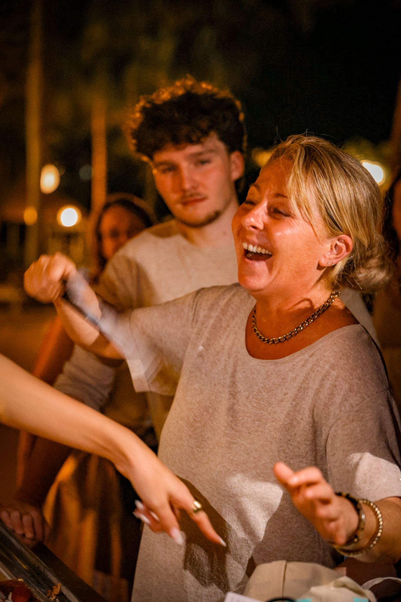 Laughing blonde woman in a grey top enjoying a lively outdoor nighttime gathering.