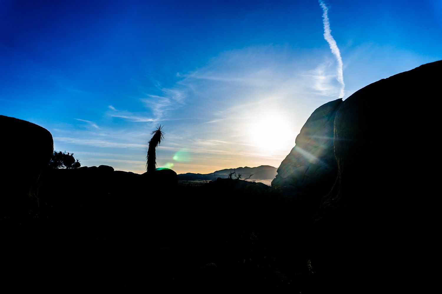 Silhouette of a person and rocks against a vibrant blue sky with sun flare at golden hour.