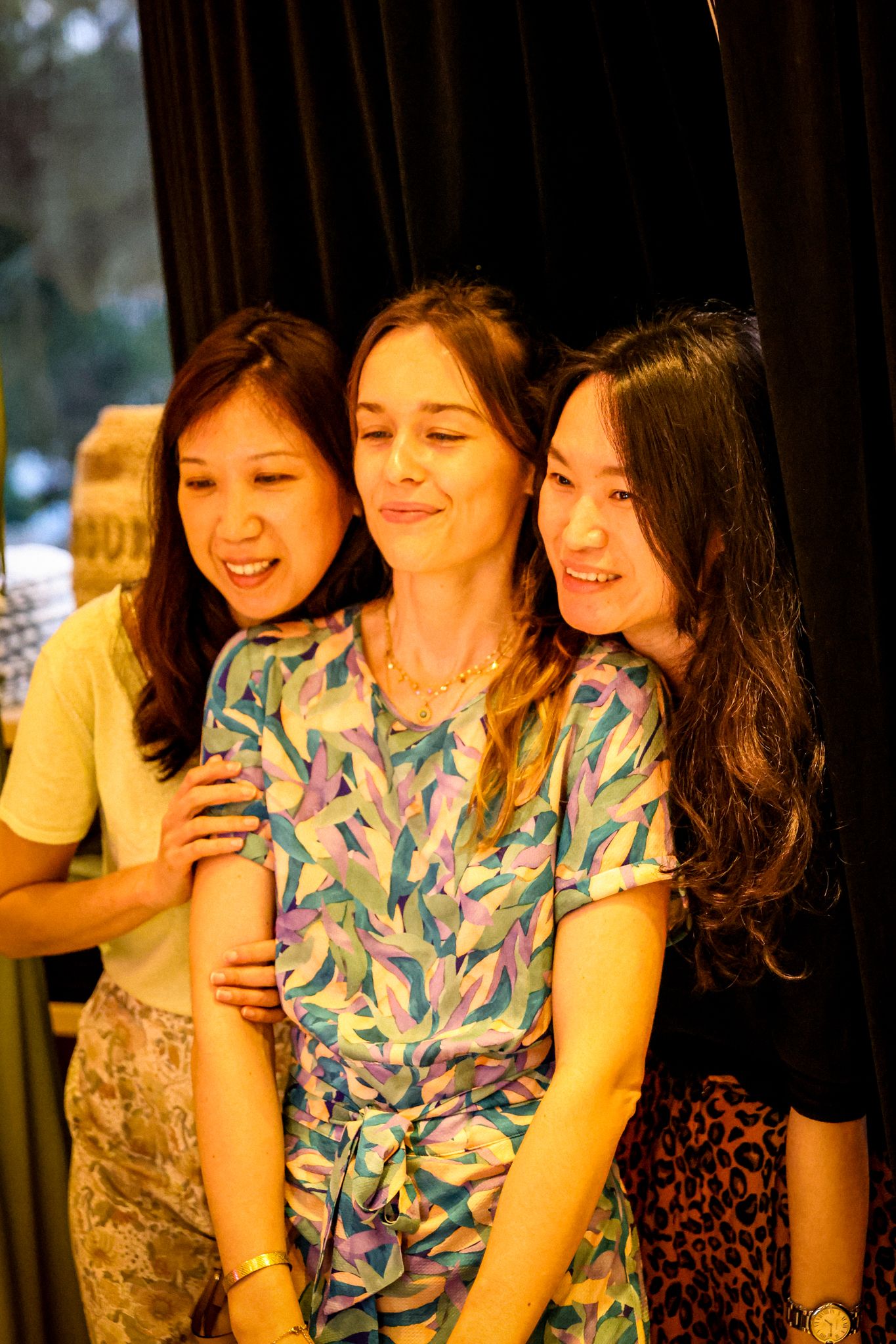 Three smiling women pose together at a social event, with the middle woman wearing a colorful floral top.