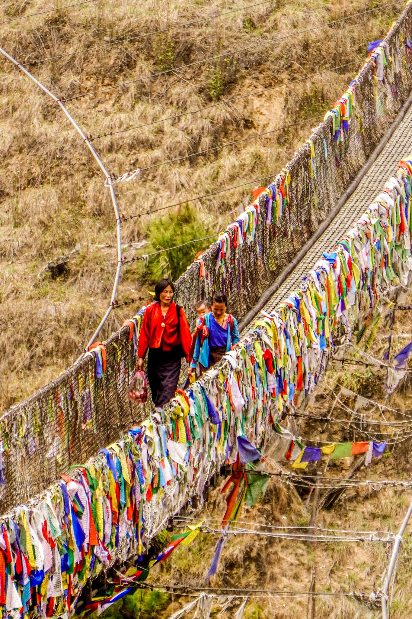 Two women walk across a vibrant suspension bridge decorated with hundreds of colorful prayer flags in Bhutan.