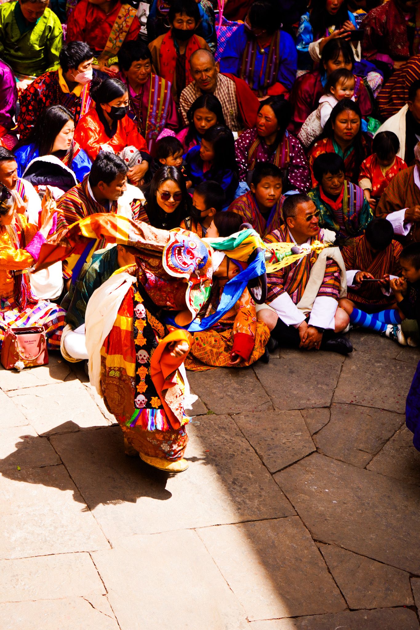 A single Bhutanese Cham dancer in ornate costume and skull mask performs sacred ritual dance before a crowd of colorful spectators.