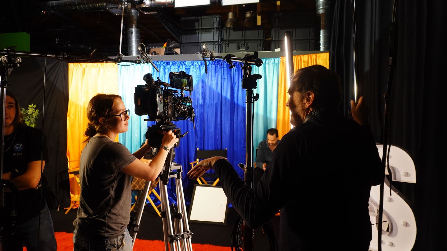 Film crew operating a professional camera on a colorful production set with blue curtain backdrop.