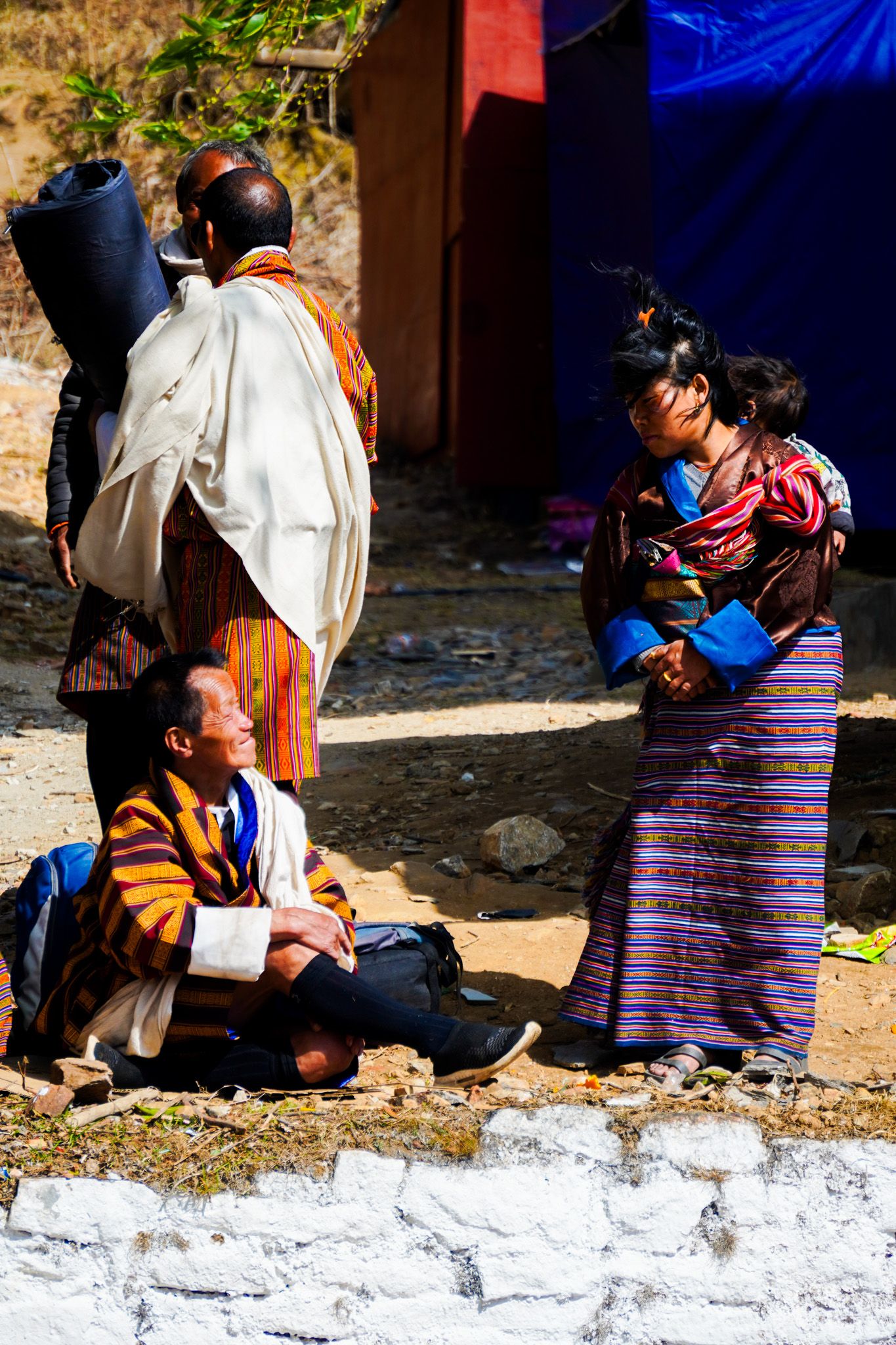 Bhutanese people in traditional dress converse outdoors near a white stone wall at a cultural event.