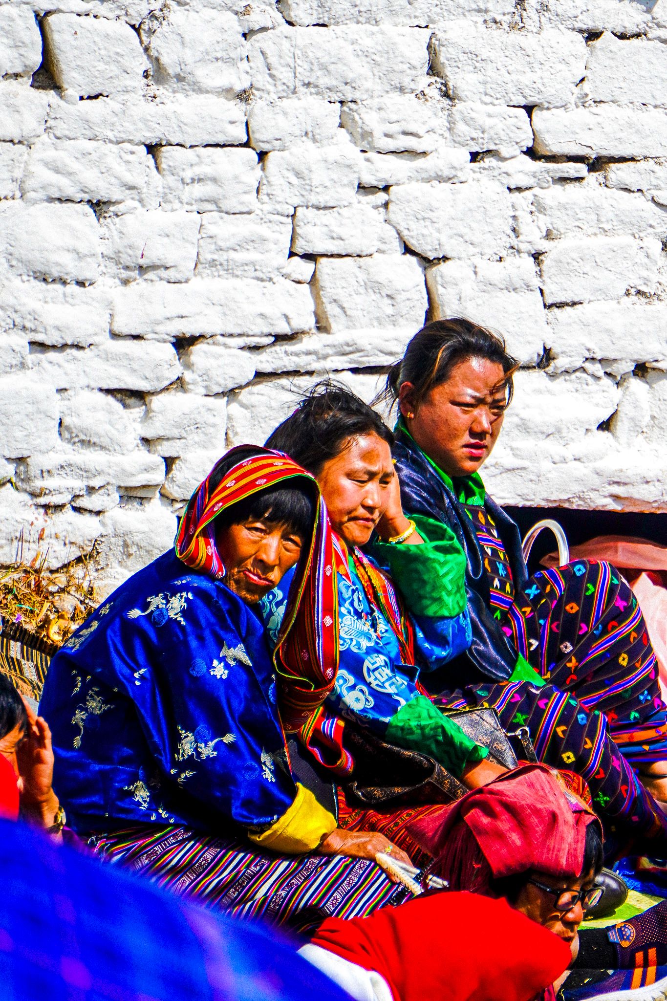 Three Bhutanese women wearing vibrant traditional clothing rest against a white stone wall outdoors.