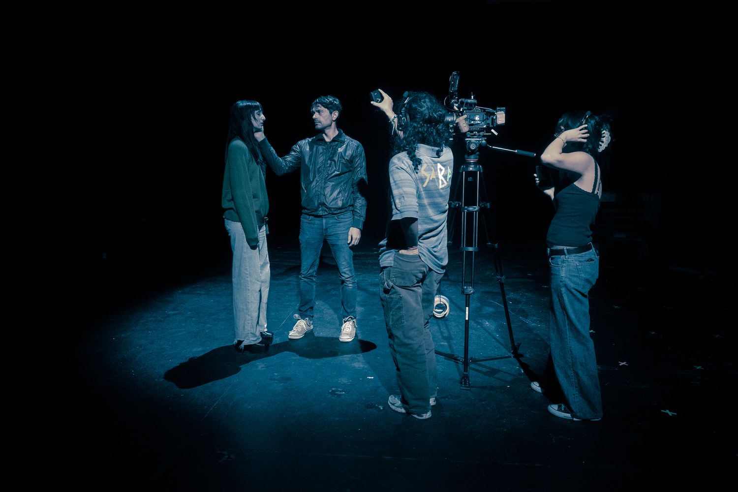 Film crew with camera operator recording actors on a dark stage under blue lighting.