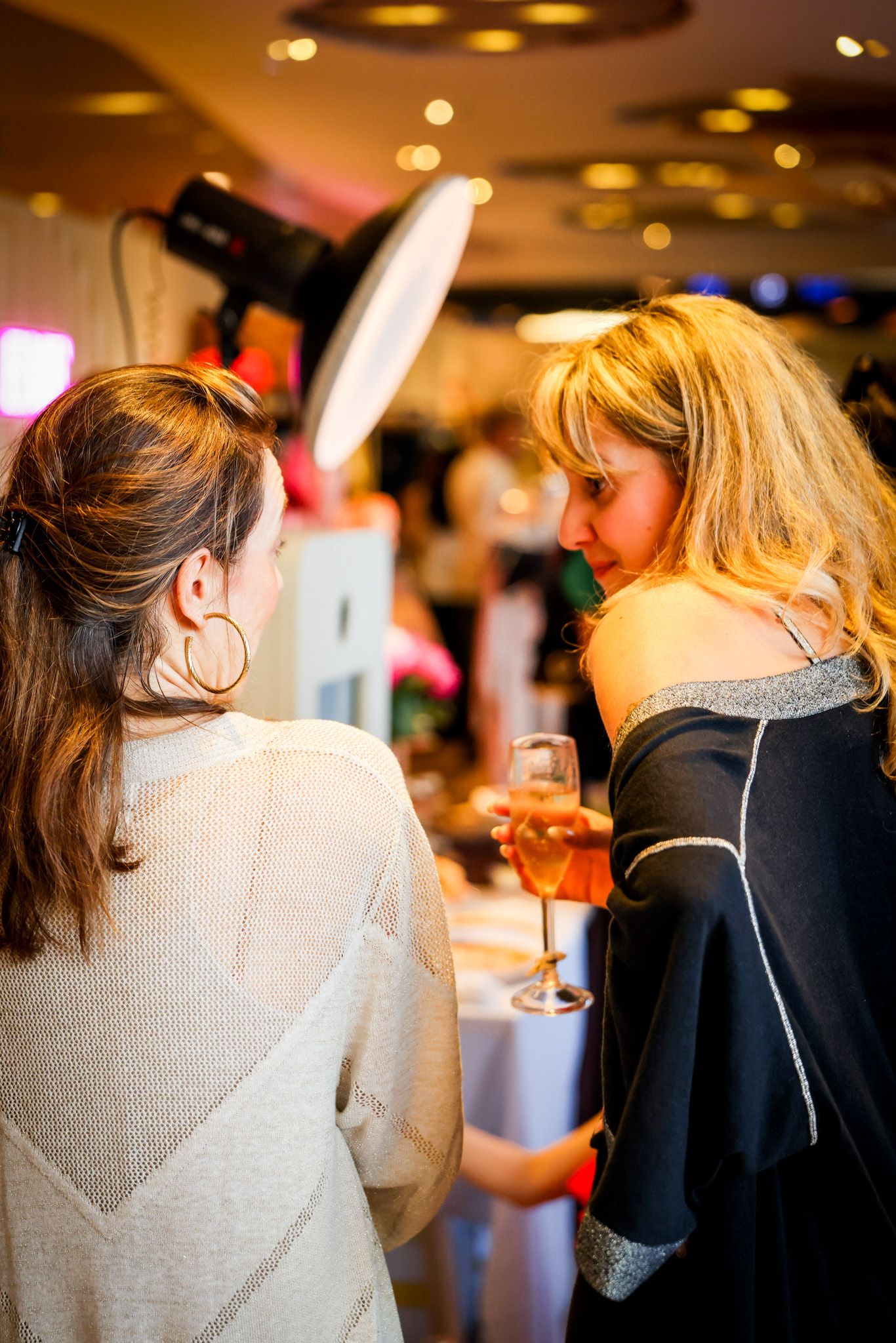 Two women chat at an elegant indoor event, one holding a glass of champagne in a warmly lit venue.