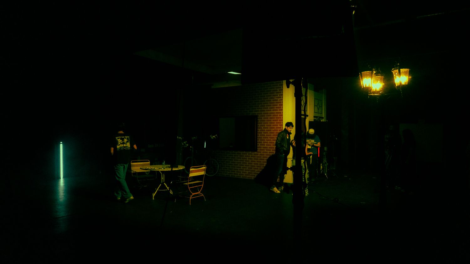 Dark film set with green ambient lighting, crew members working around a table and lamp post.