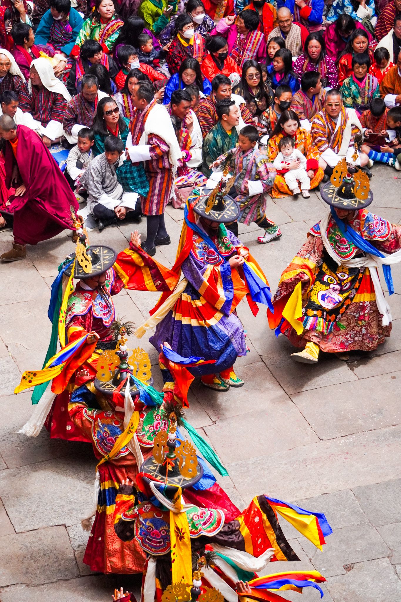 Multiple elaborately costumed Cham dancers perform sacred Bhutanese Tsechu festival masked dance before a large colorful audience.